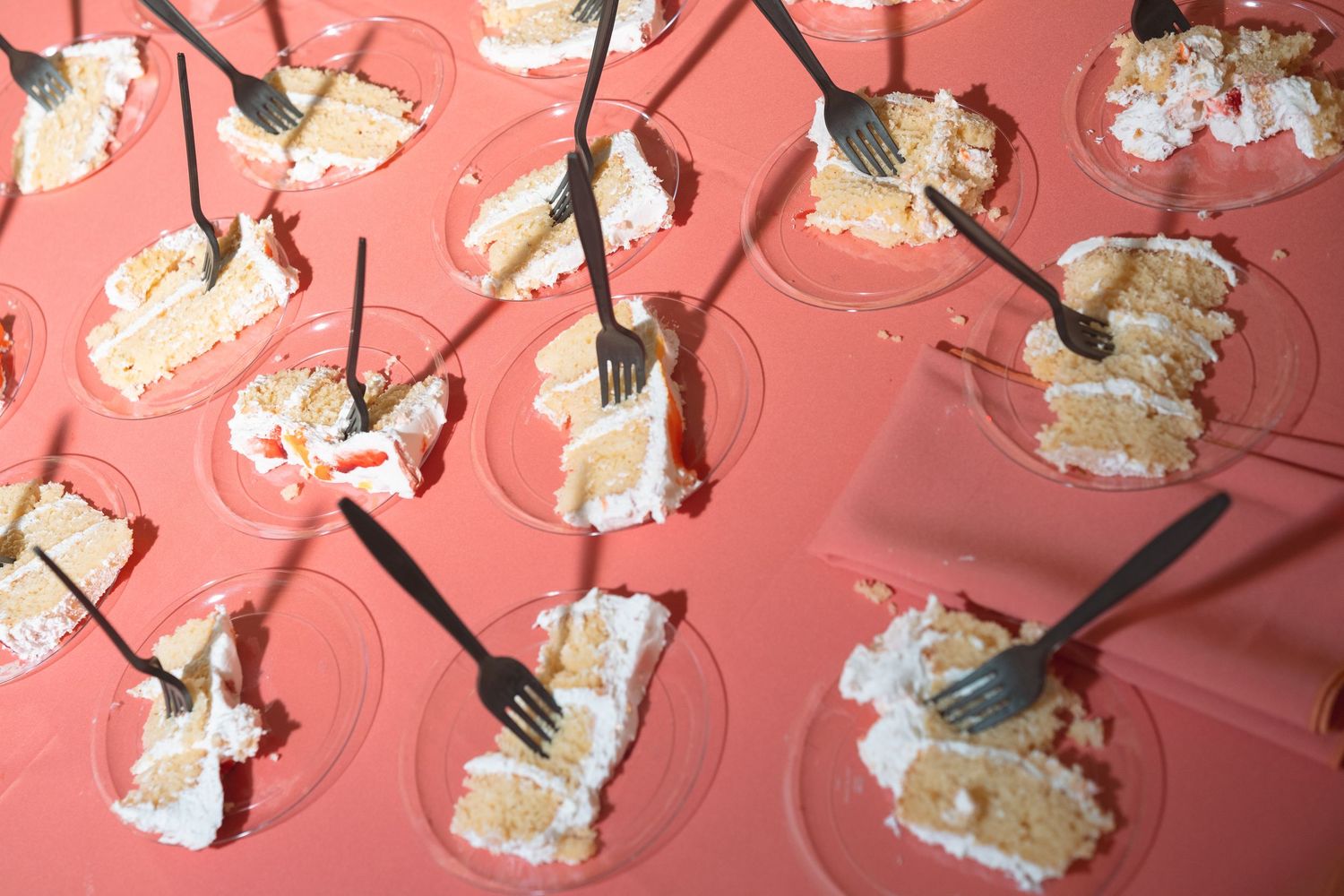 Multiple plates of white frosted cake slices with forks on a pink tablecloth at a catered event.