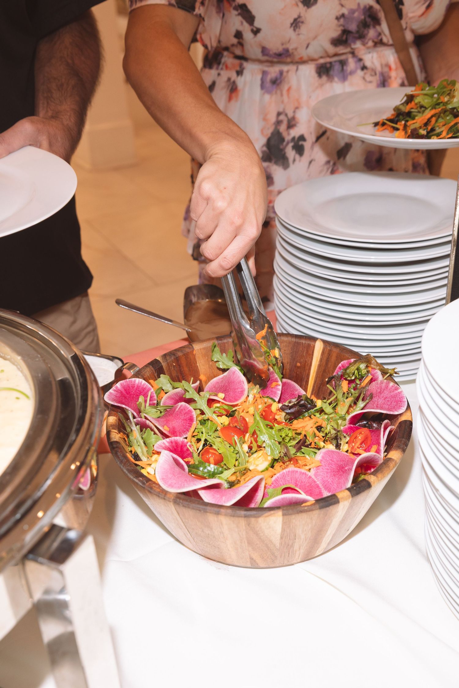 Person serving a colorful watermelon radish salad with tongs at a buffet table with stacked white plates nearby.