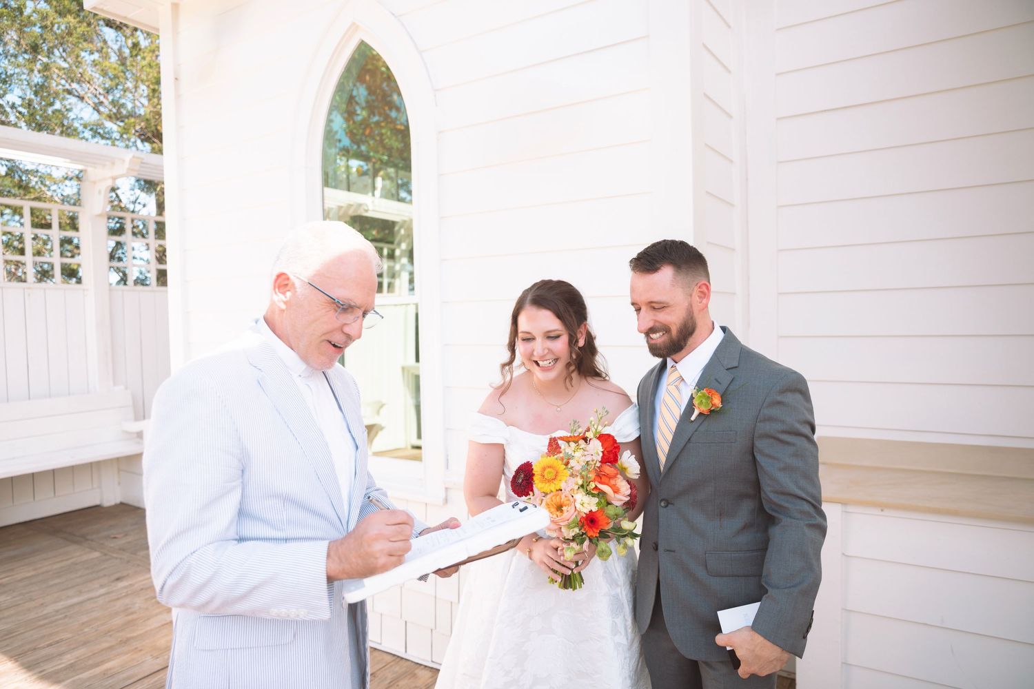 Bride and groom smiling with officiant during outdoor wedding ceremony at white chapel, bride holding colorful bouquet.