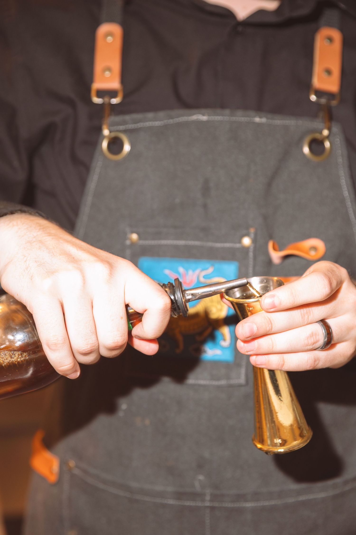 Bartender in gray apron pouring spirits into a brass jigger at a wedding bar.