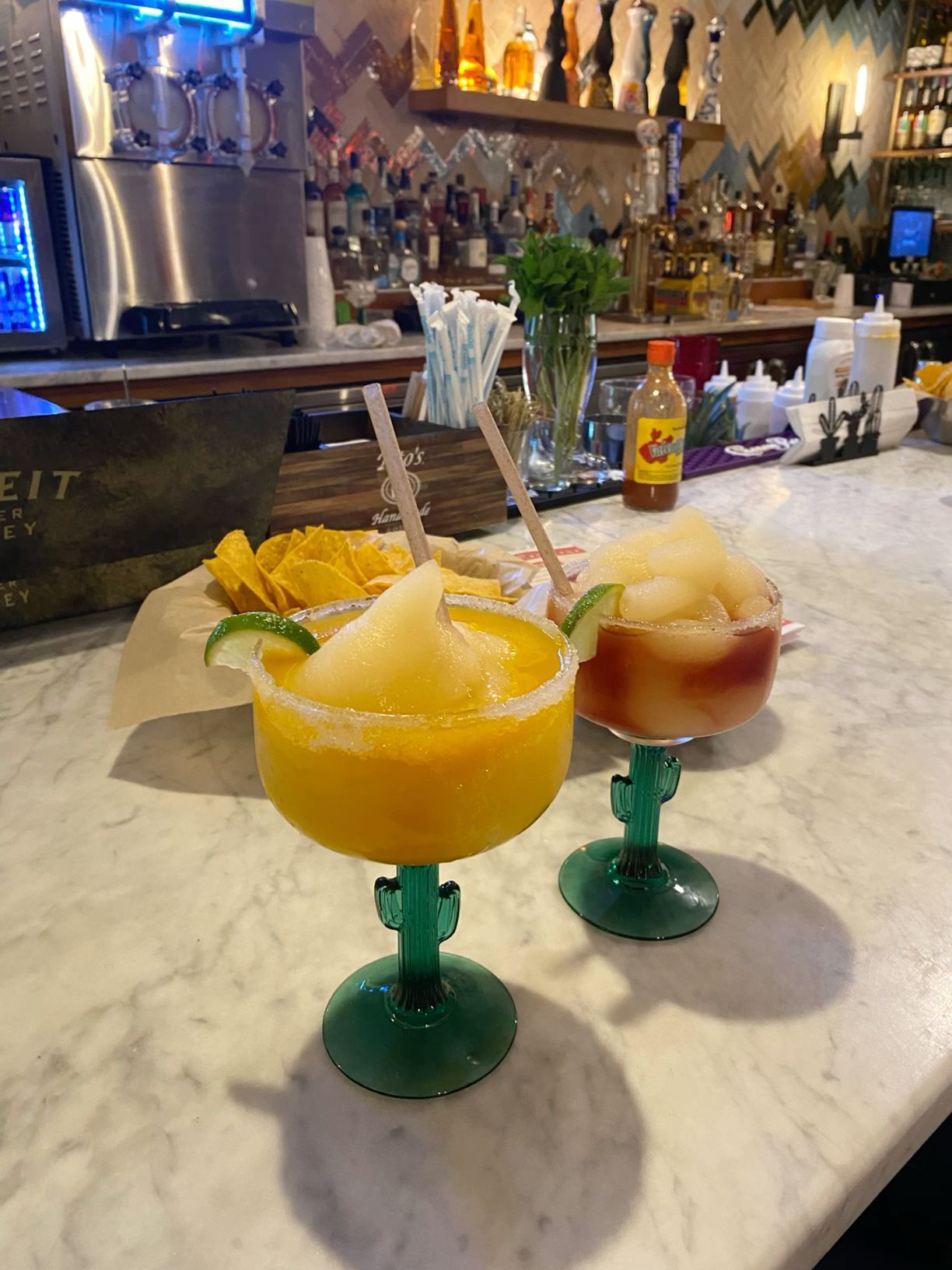 Colorful frozen margaritas in cactus glasses on marble bar counter with tropical garnishes.