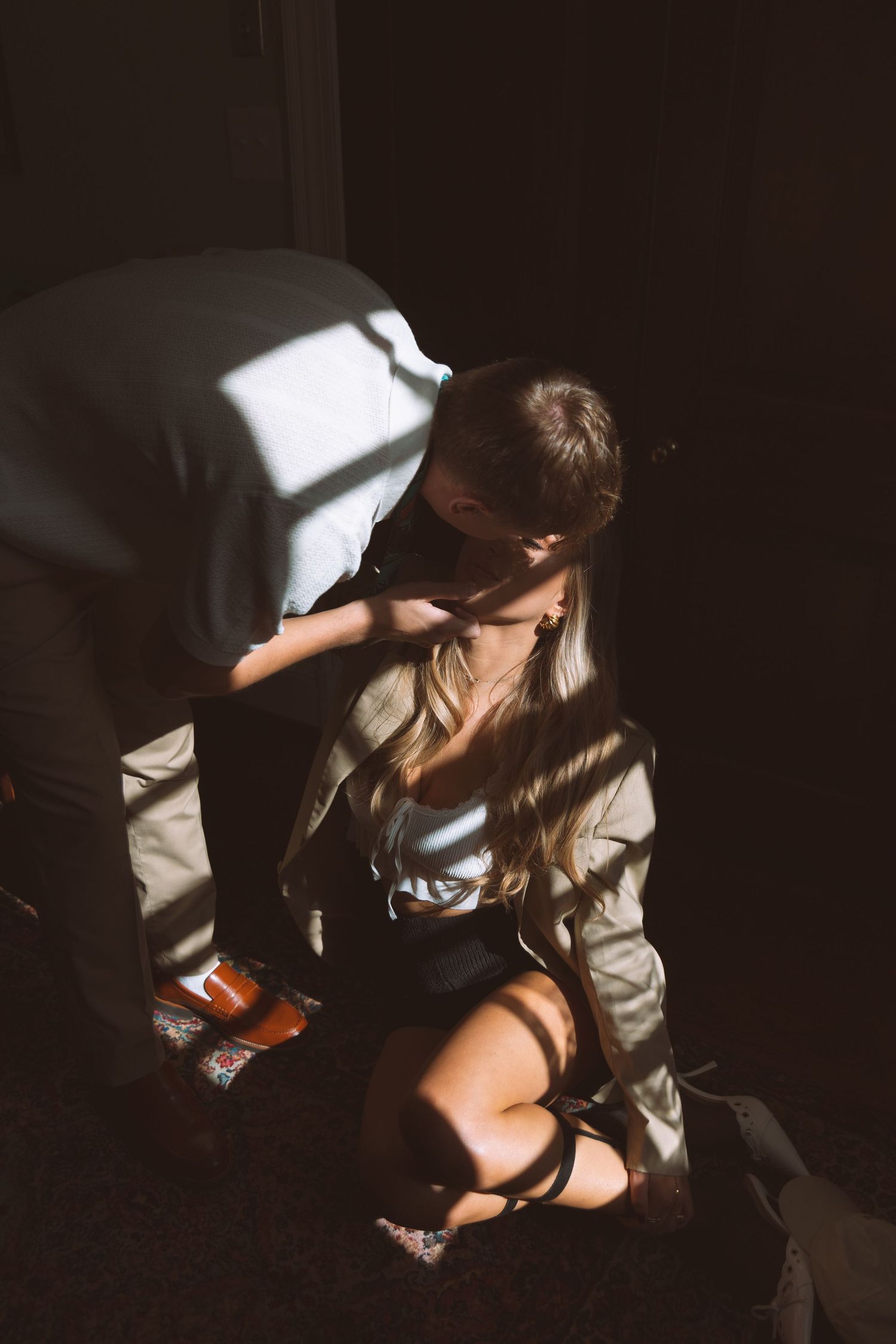 Groom helping bride with shoes in dramatic sunlit doorway shadow.