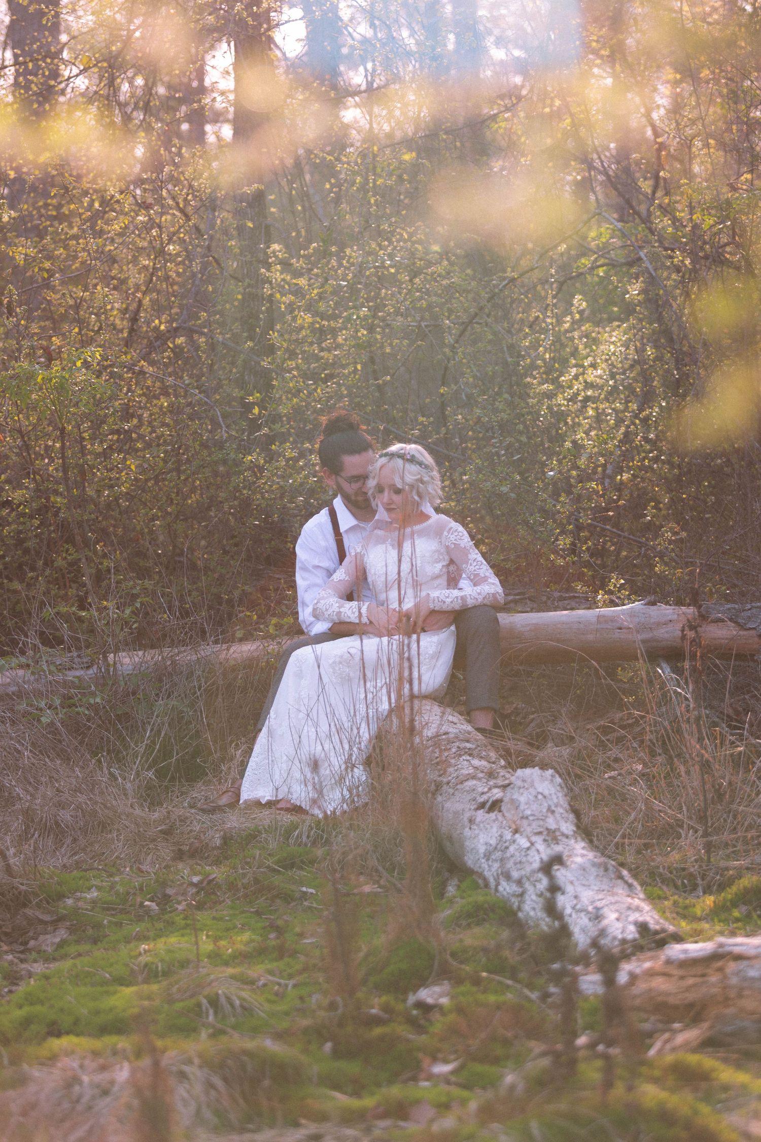 Bride and groom sit intimately on a fallen log in a sunlit forest setting.