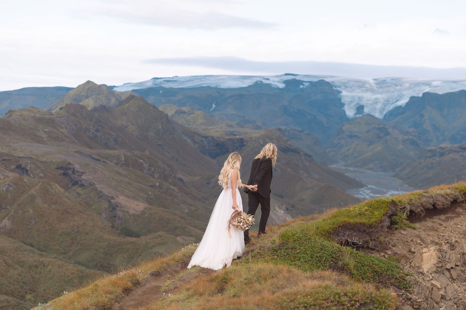 Bride and groom hold hands on a mountain cliff overlooking a glacier and valley in Iceland.