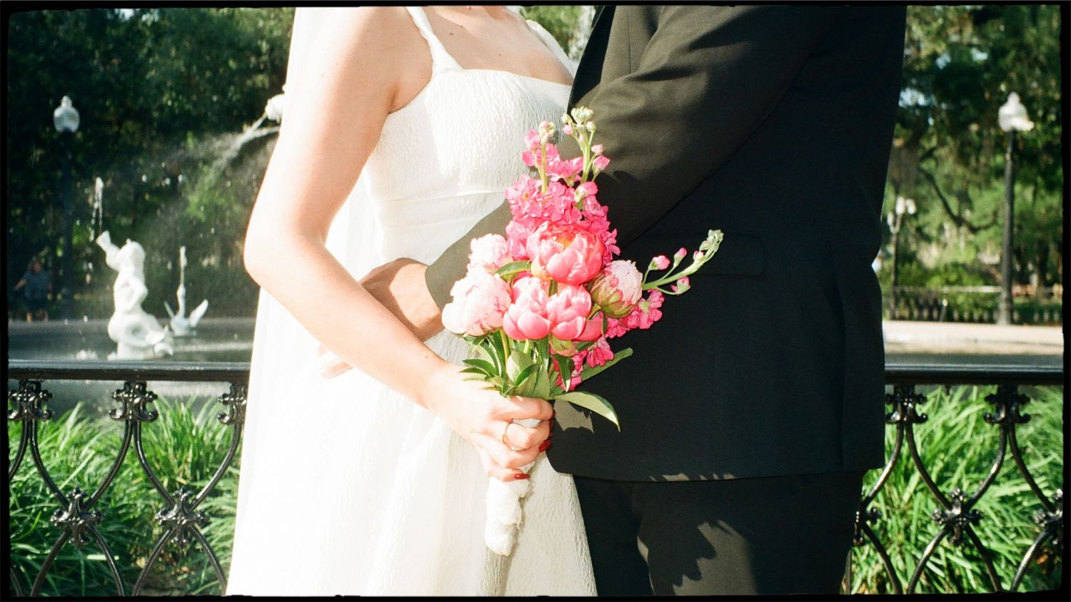 Bride holds pink peony bouquet beside groom in black suit with garden fountain backdrop at wedding.