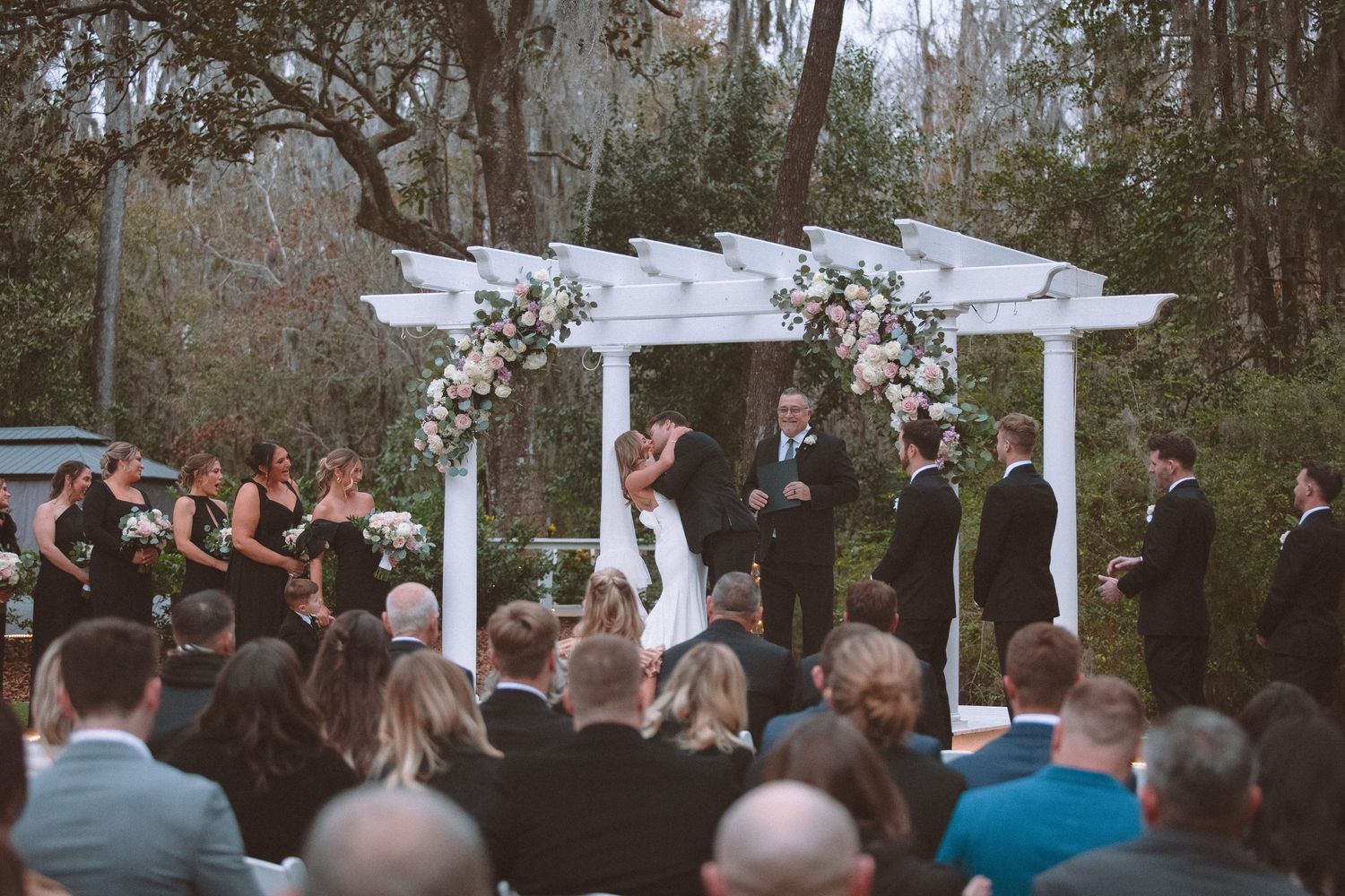 Couple shares first kiss under a white floral pergola arch surrounded by bridesmaids and groomsmen.