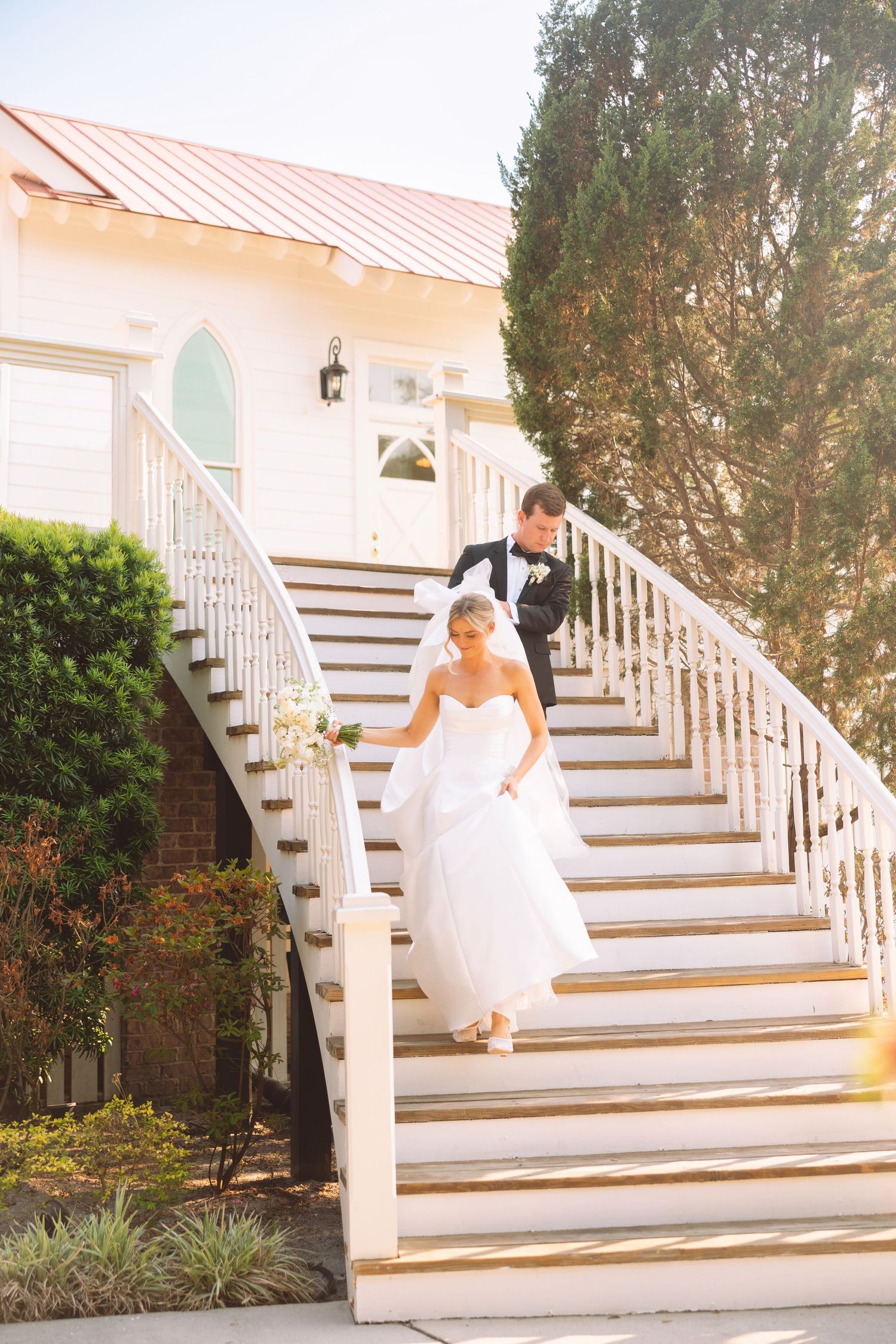 Bride and groom descend grand white staircase at sunlit wedding venue.