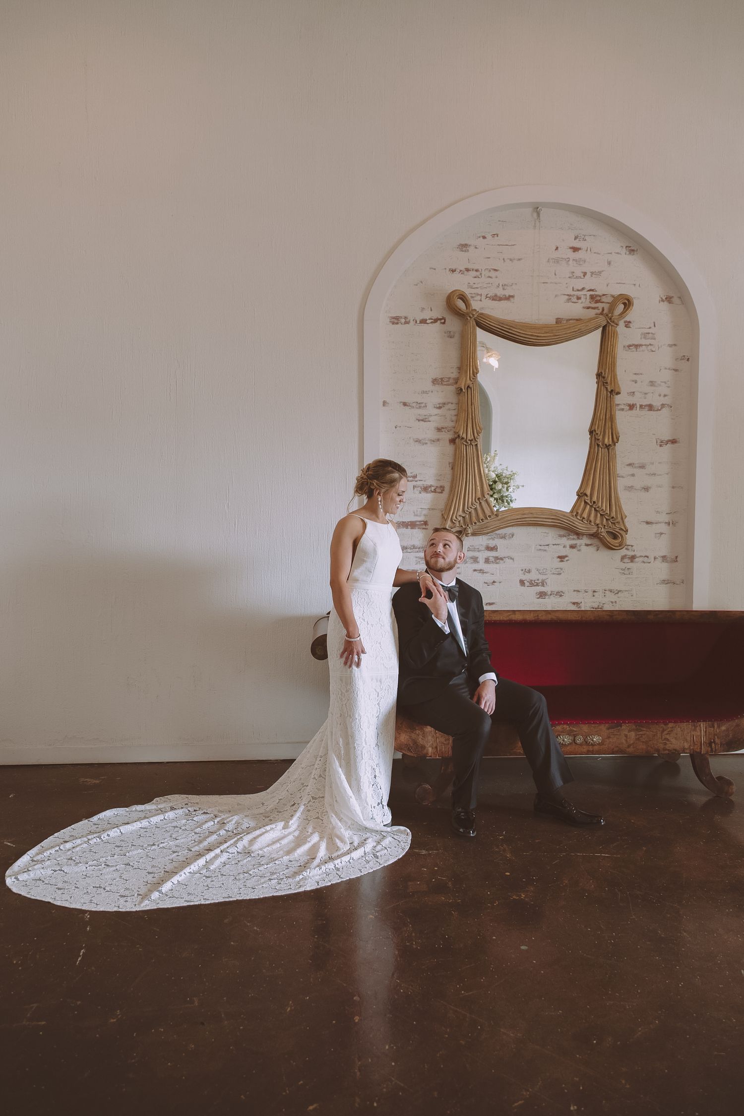 Bride in long lace train gown stands beside seated groom in black suit near arched mirror.