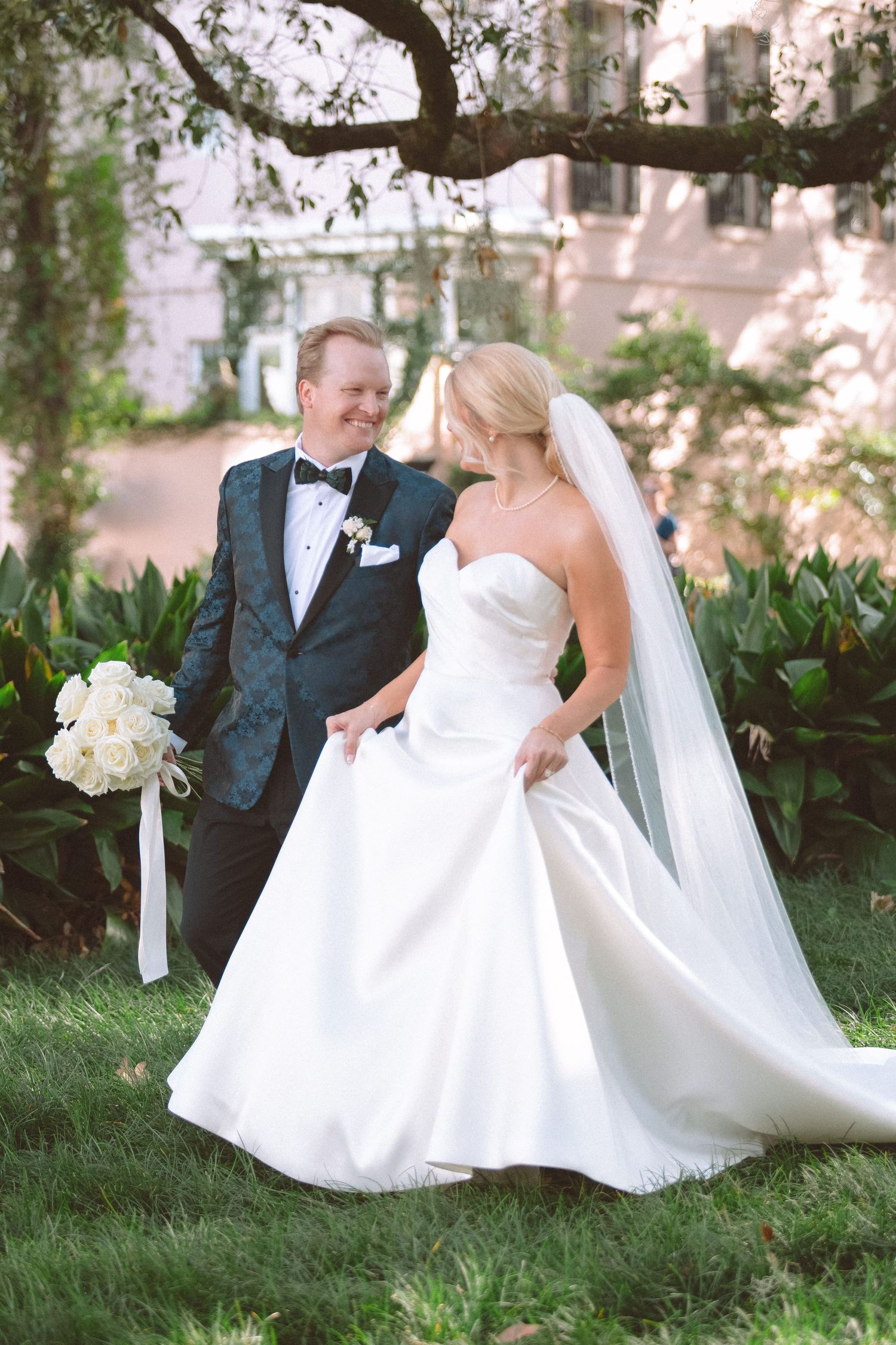 Bride in strapless satin ballgown with veil and groom in navy tuxedo sharing a romantic moment outdoors.