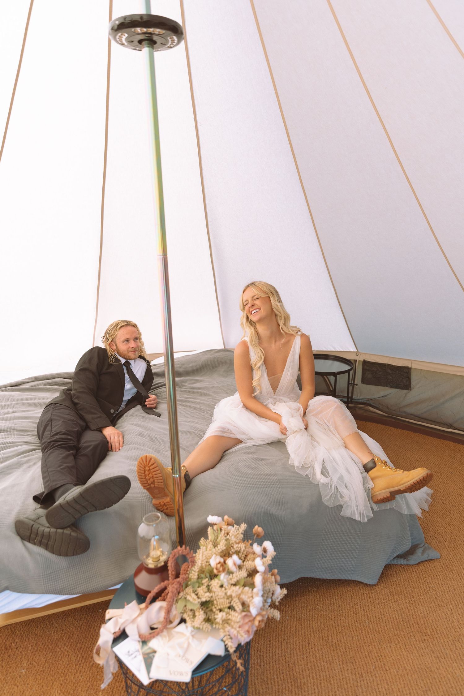 A bride in a white dress and groom in a suit relax inside a boho tipi tent during their wedding celebration.