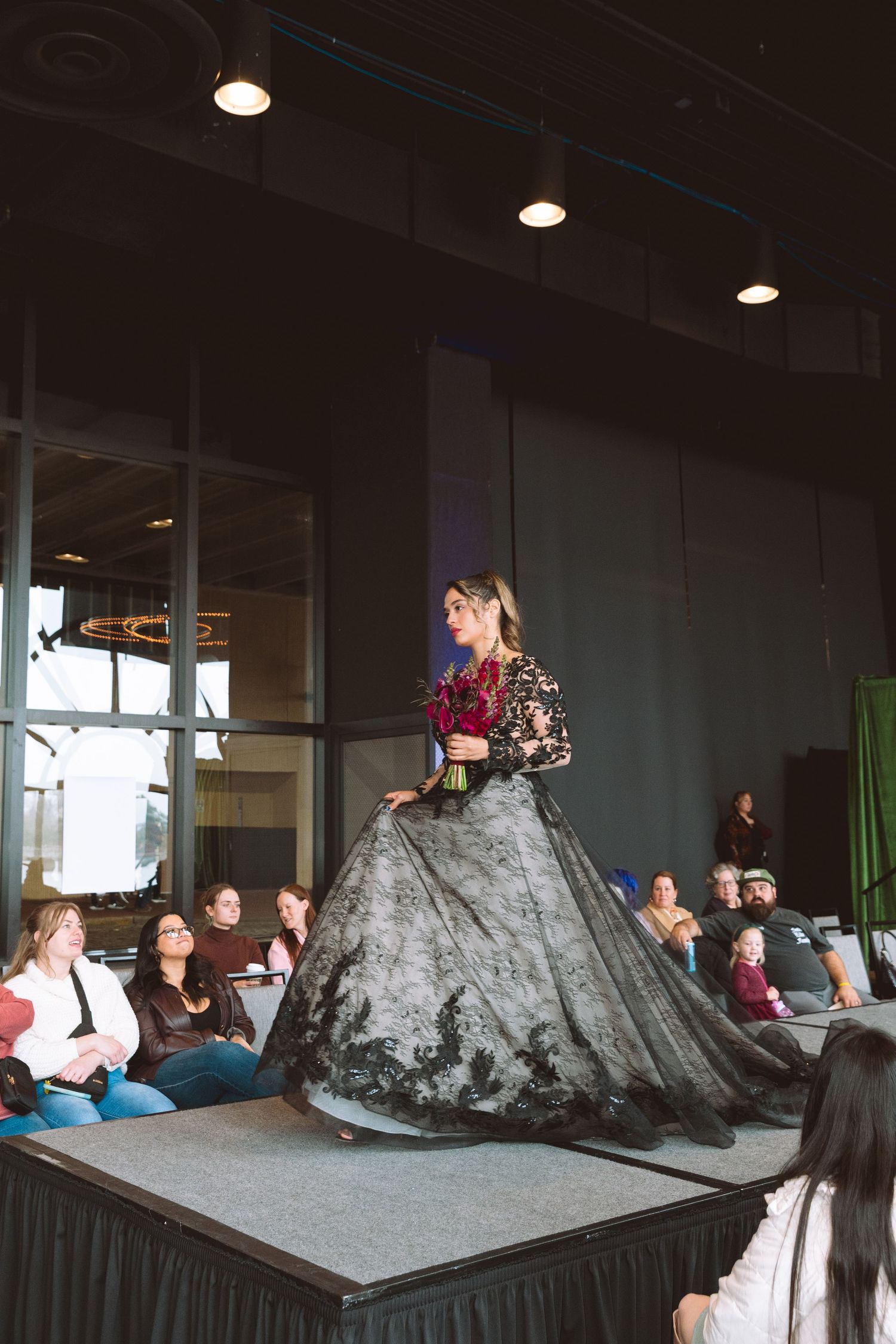 Model walks runway in stunning black lace ballgown with red floral bodice at indoor fashion show.