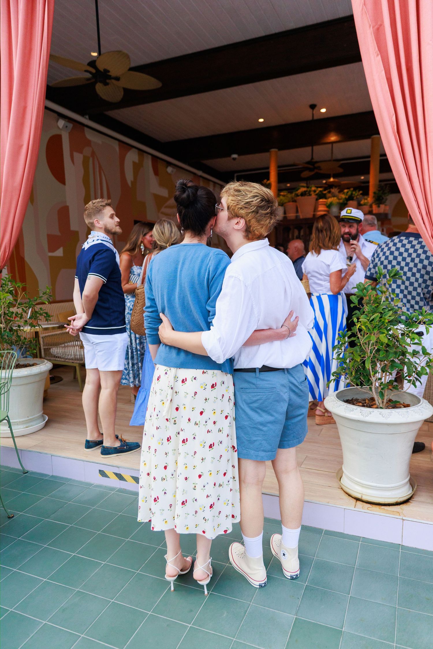 Couple sharing a kiss at a colorful outdoor party venue with guests mingling in the background.
