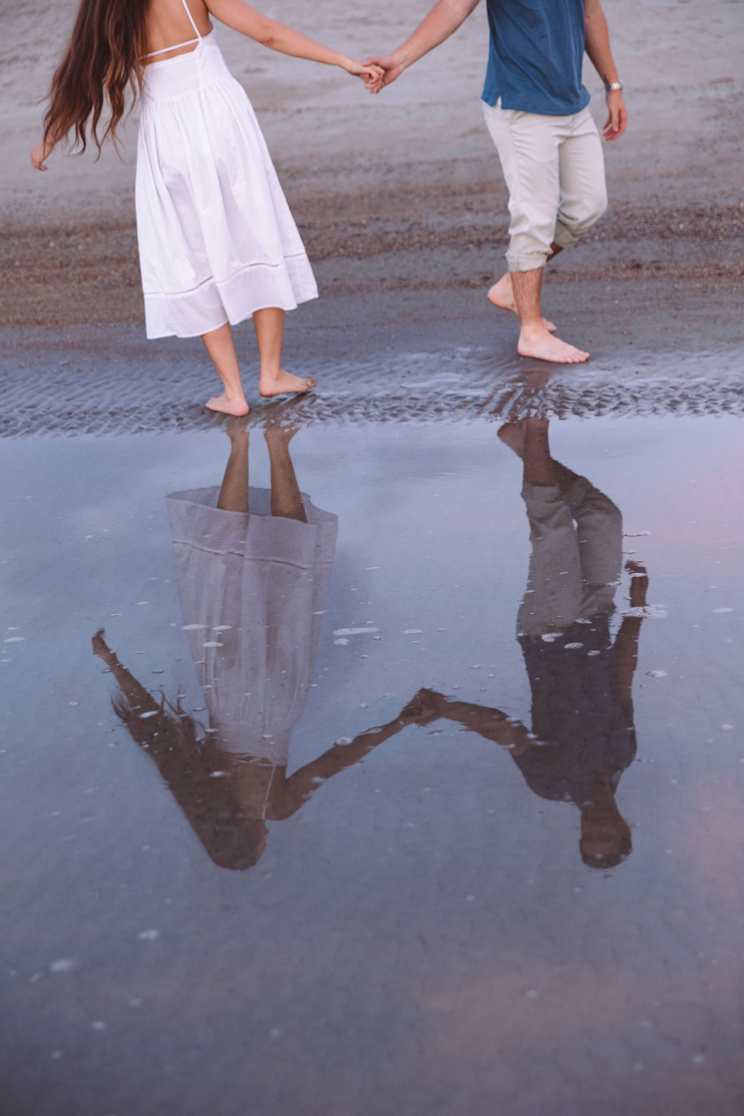 Barefoot couple holding hands on wet beach sand with romantic reflection visible in the water below.