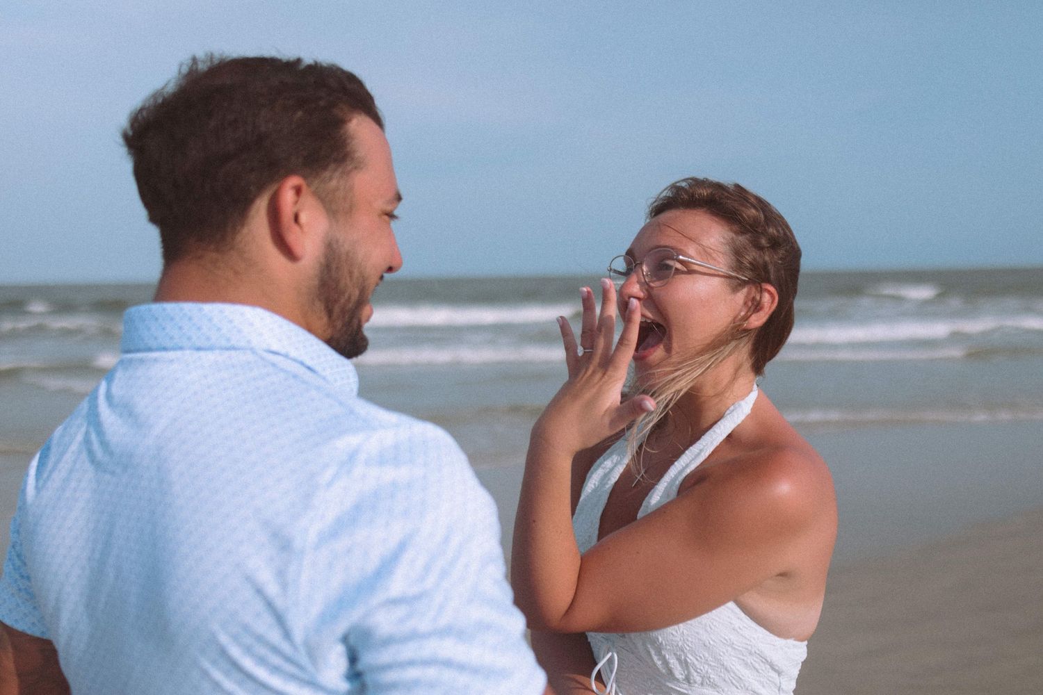 Woman reacting with surprise and joy to a marriage proposal on a sunny beach.
