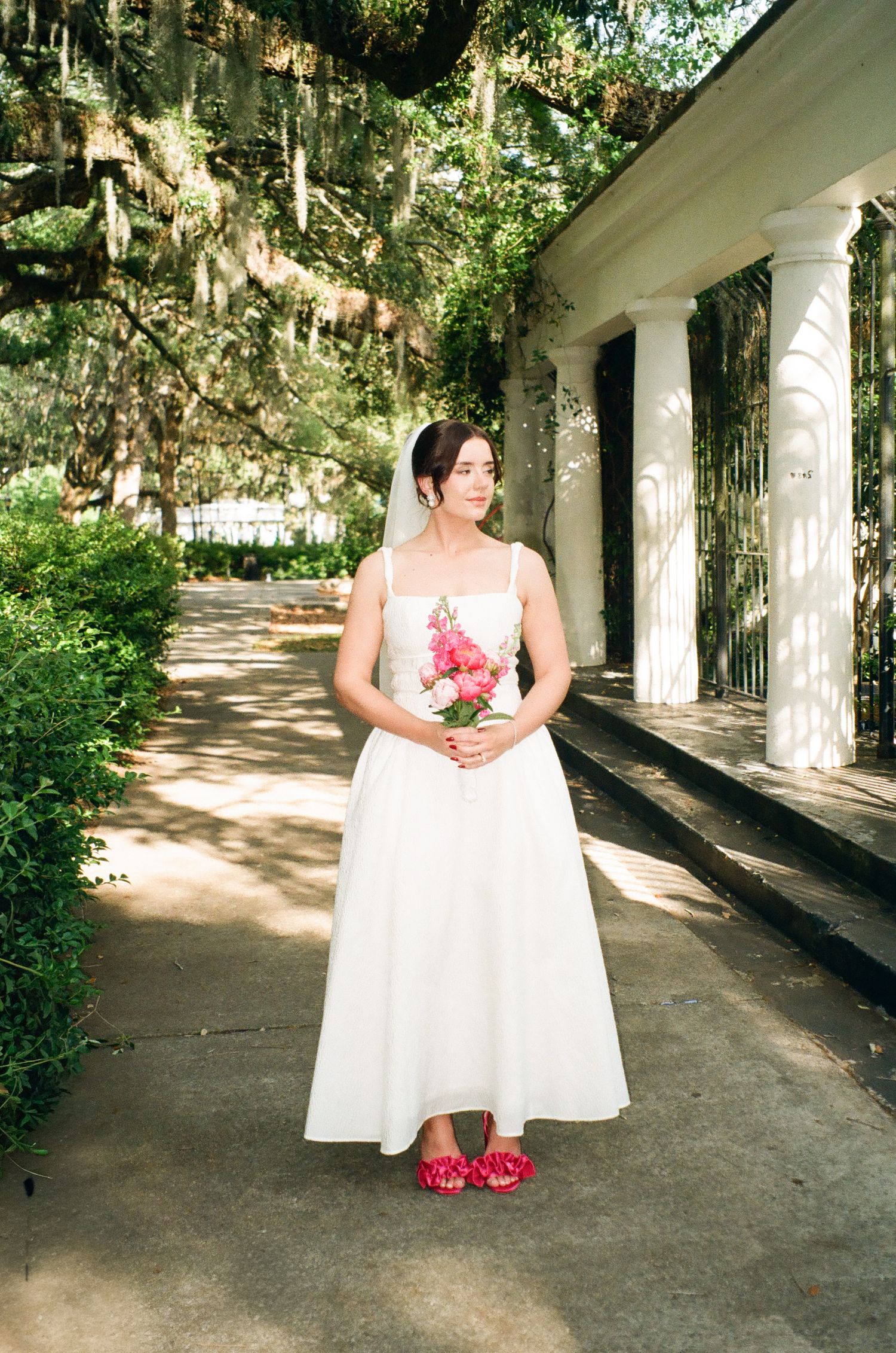 A bride in a white midi dress and red shoes holds a pink bouquet outdoors near white columns and mossy trees.