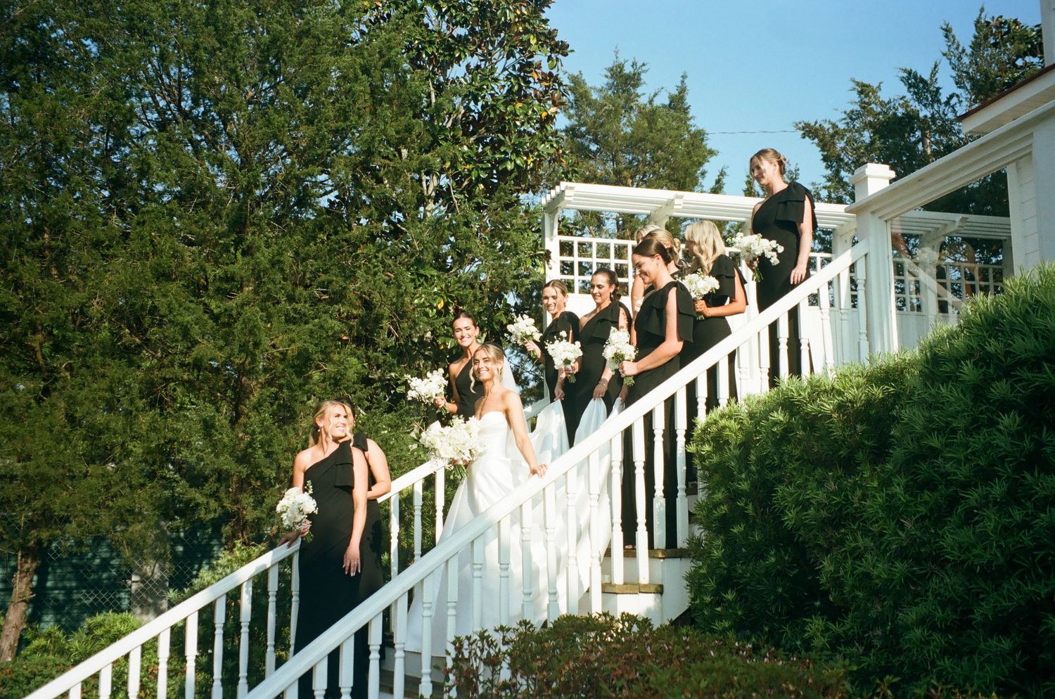 Bridal party in black gowns descending white staircase at outdoor wedding venue.