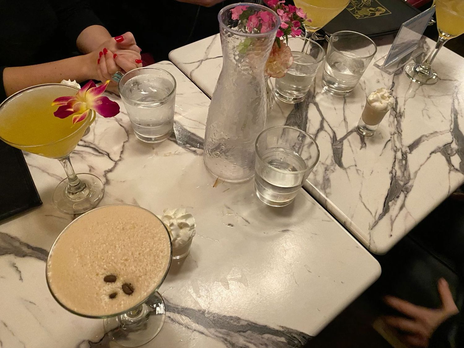 Colorful cocktails on marble table including espresso martini and yellow drink with tropical flower garnish.