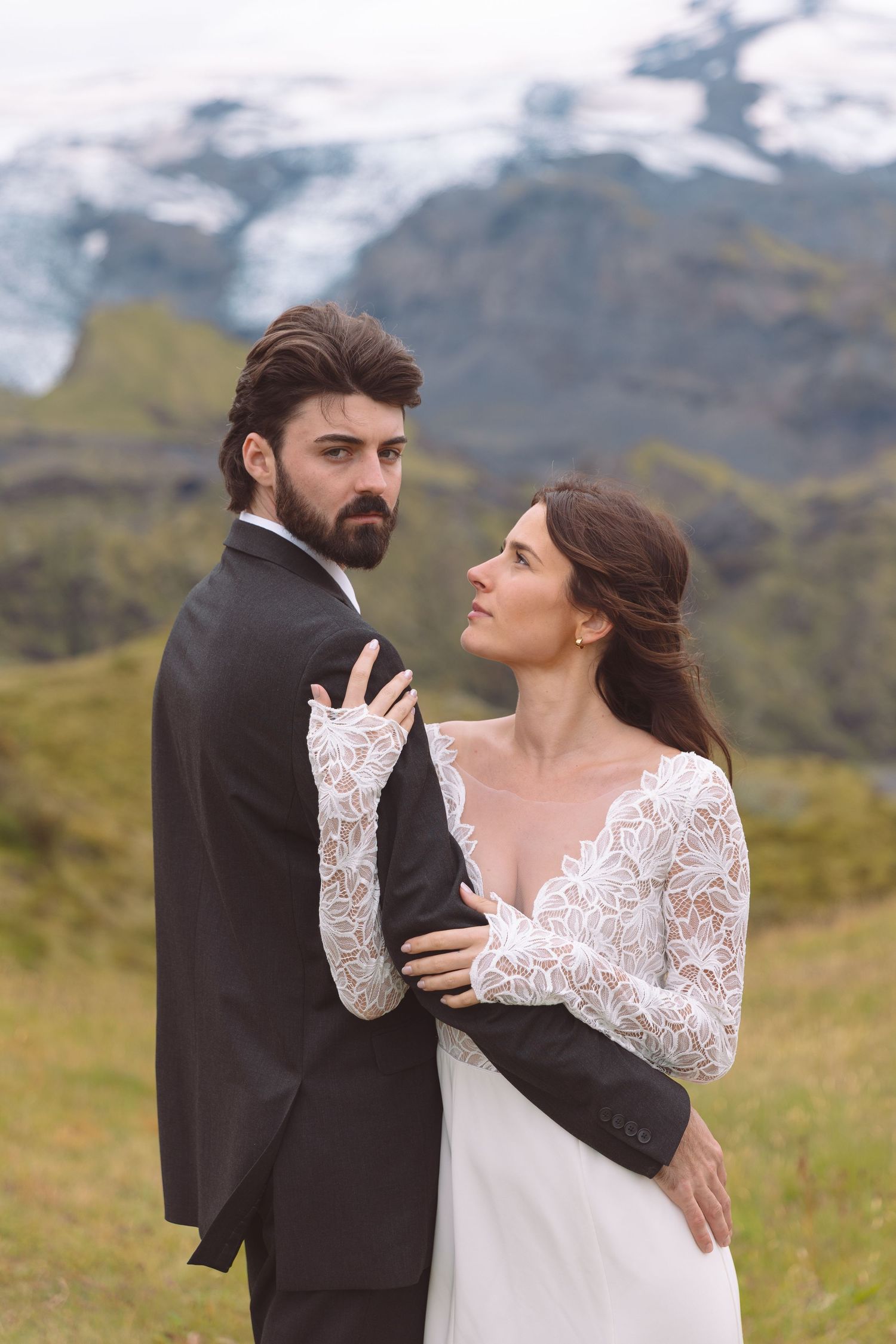 Wedding couple embracing outdoors, bride in lace dress gazes up at bearded groom near glacier.