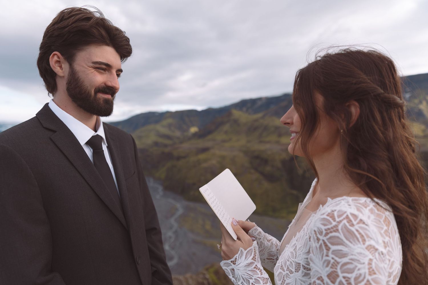 Bearded groom in black suit smiles as bride reads handwritten vows during Iceland mountain elopement.