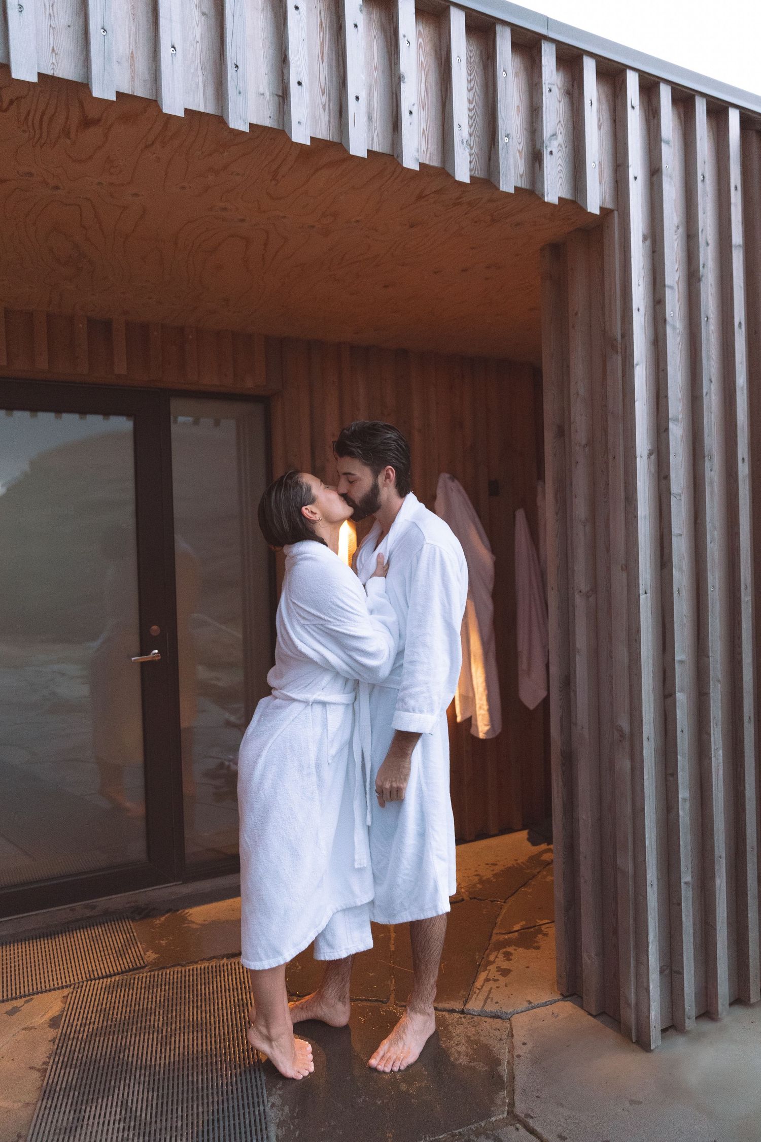 Romantic couple in white bathrobes kissing outside wooden spa facility exterior.