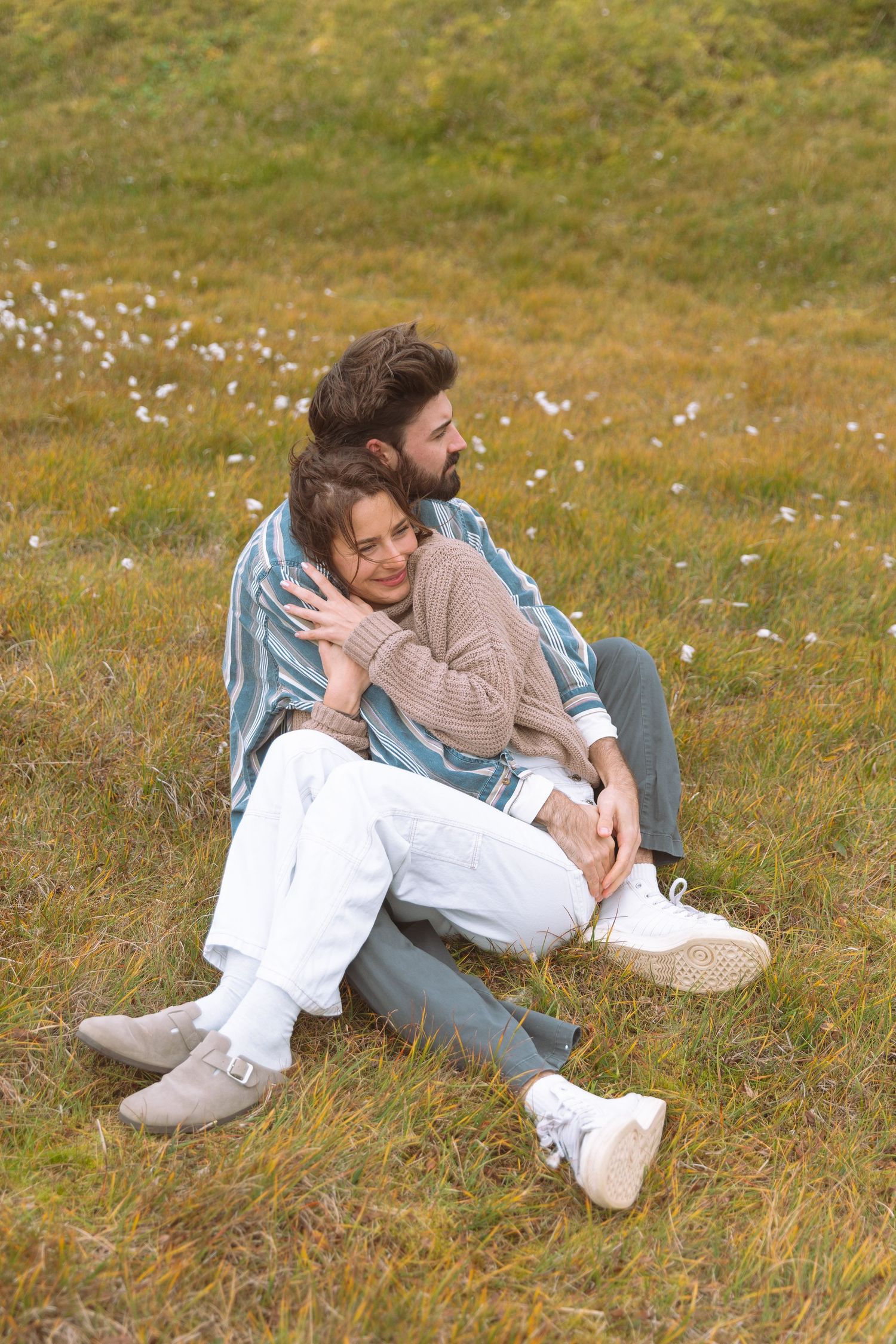 Couple sitting on grassy meadow, man embracing woman from behind, both smiling outdoors.