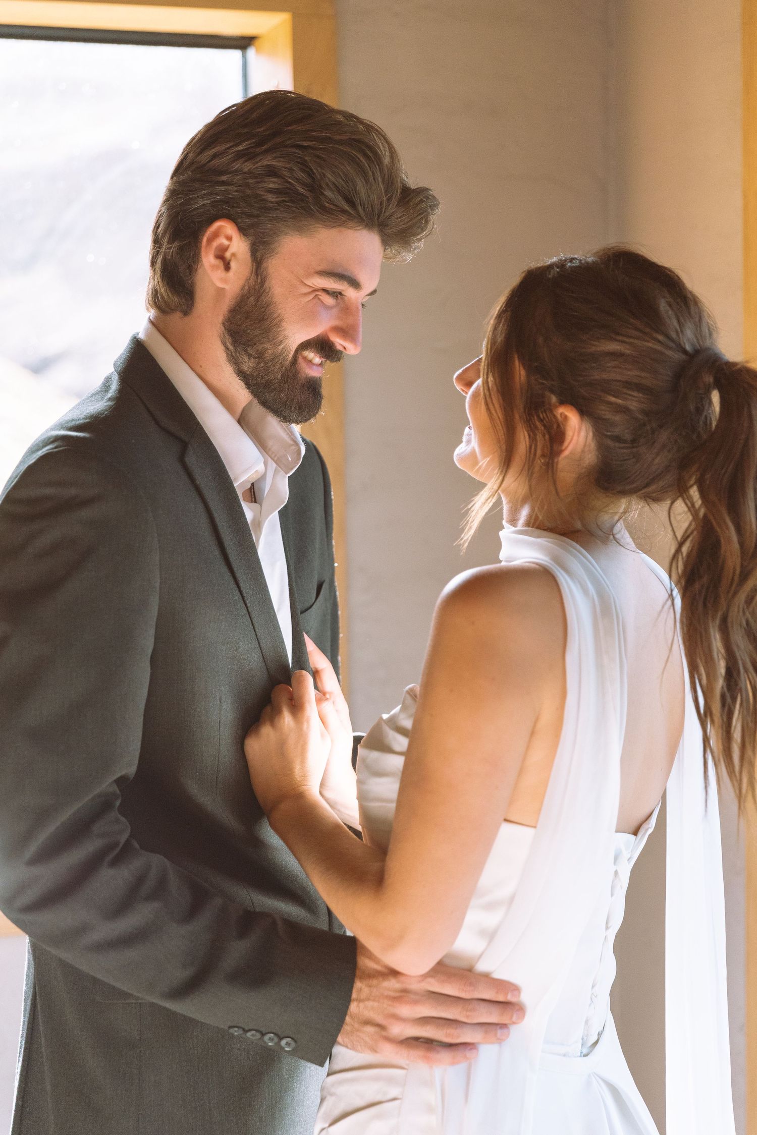 Smiling groom gazing at bride while sharing a warm embrace in natural window light.