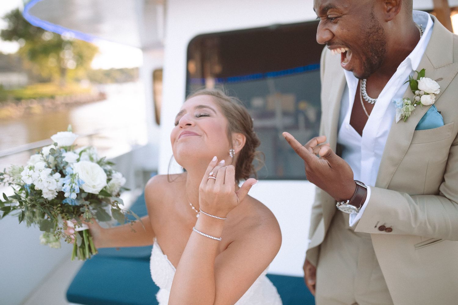 Joyful interracial bride and groom laughing together on a boat holding a white floral bouquet.