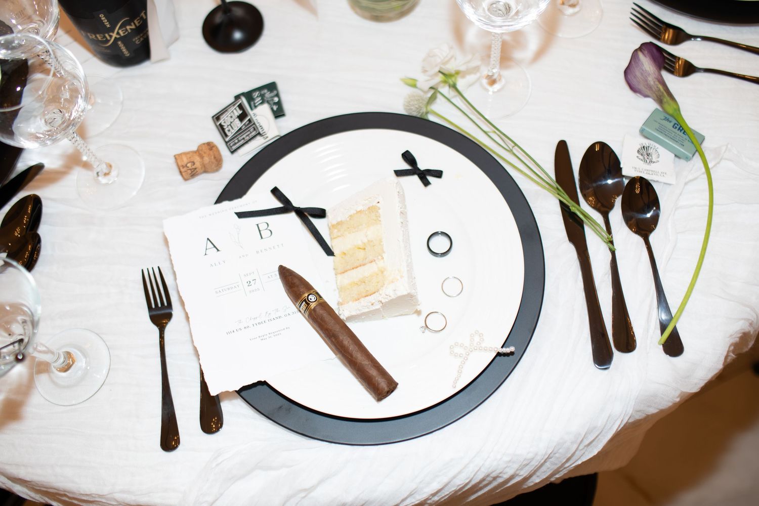 Elegant black and white wedding table setting with cigar, menu card, dark cutlery, and floral accents.
