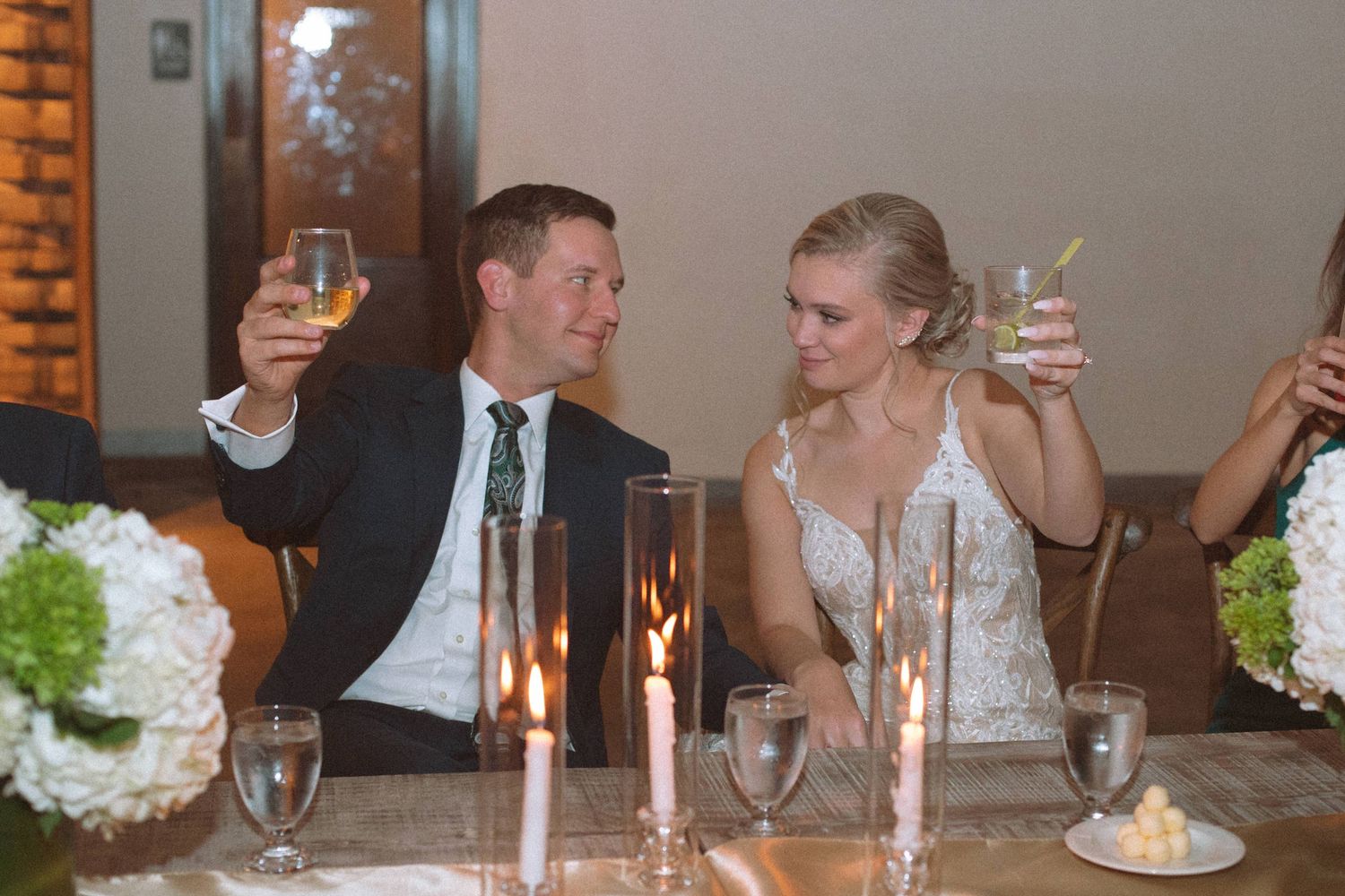 Bride and groom smile while raising champagne glasses in candlelit toast at wedding reception.