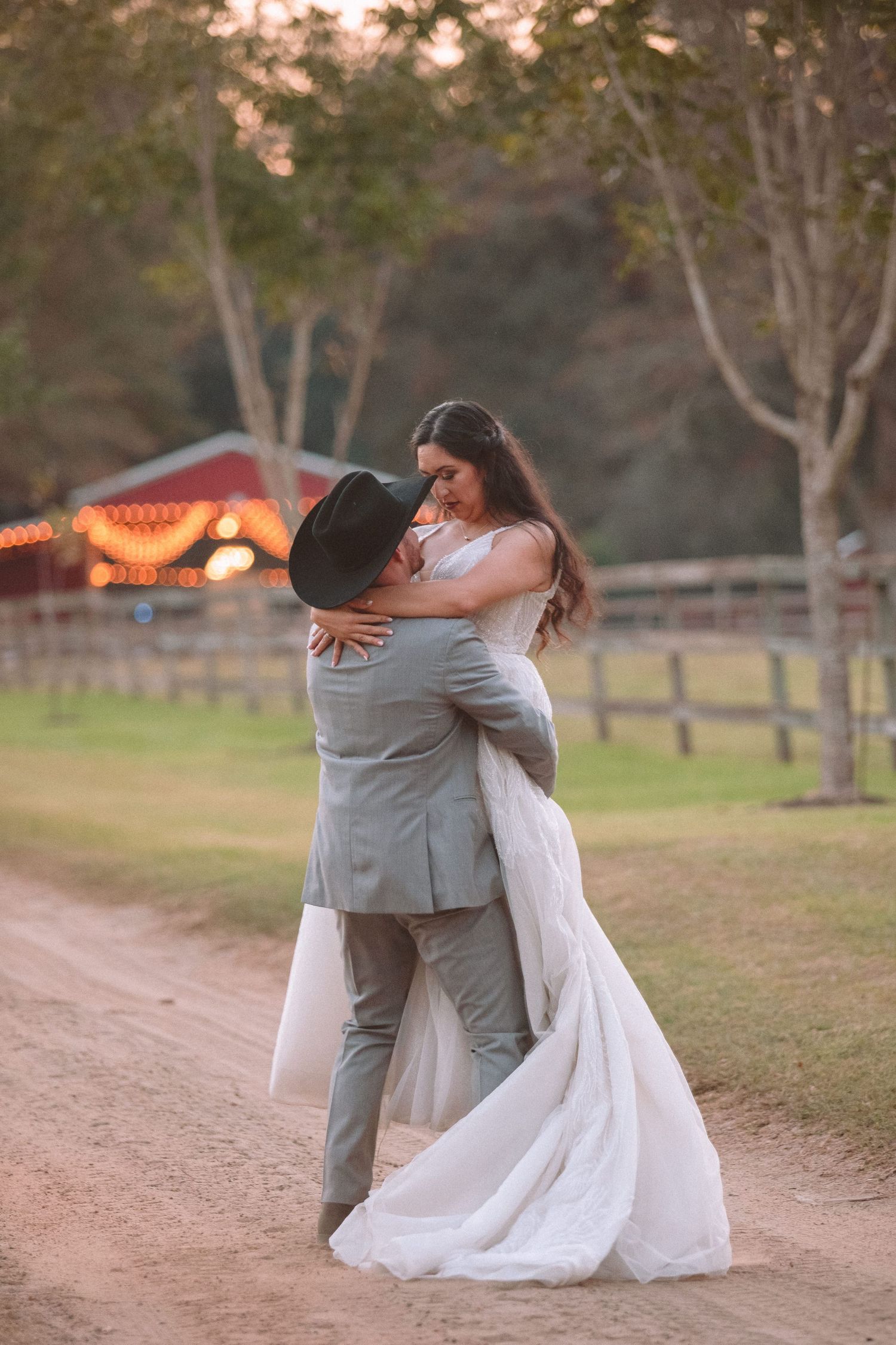 Groom in black cowboy hat lifts bride in white gown on dirt path at sunset.