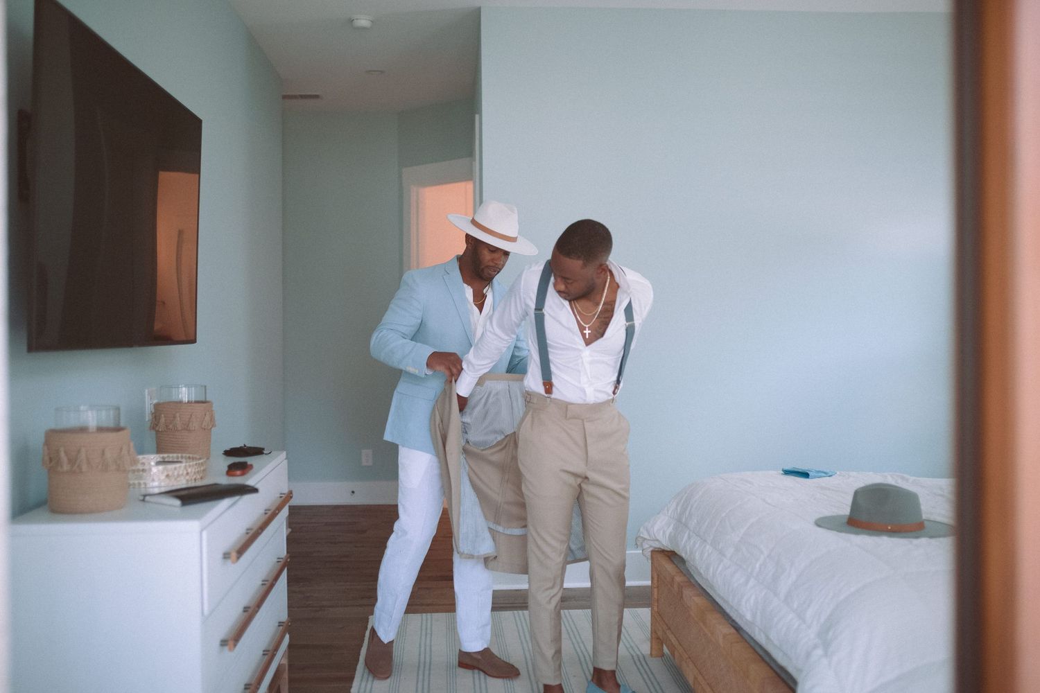 Two men in beige suits getting ready together in a light blue hotel room.