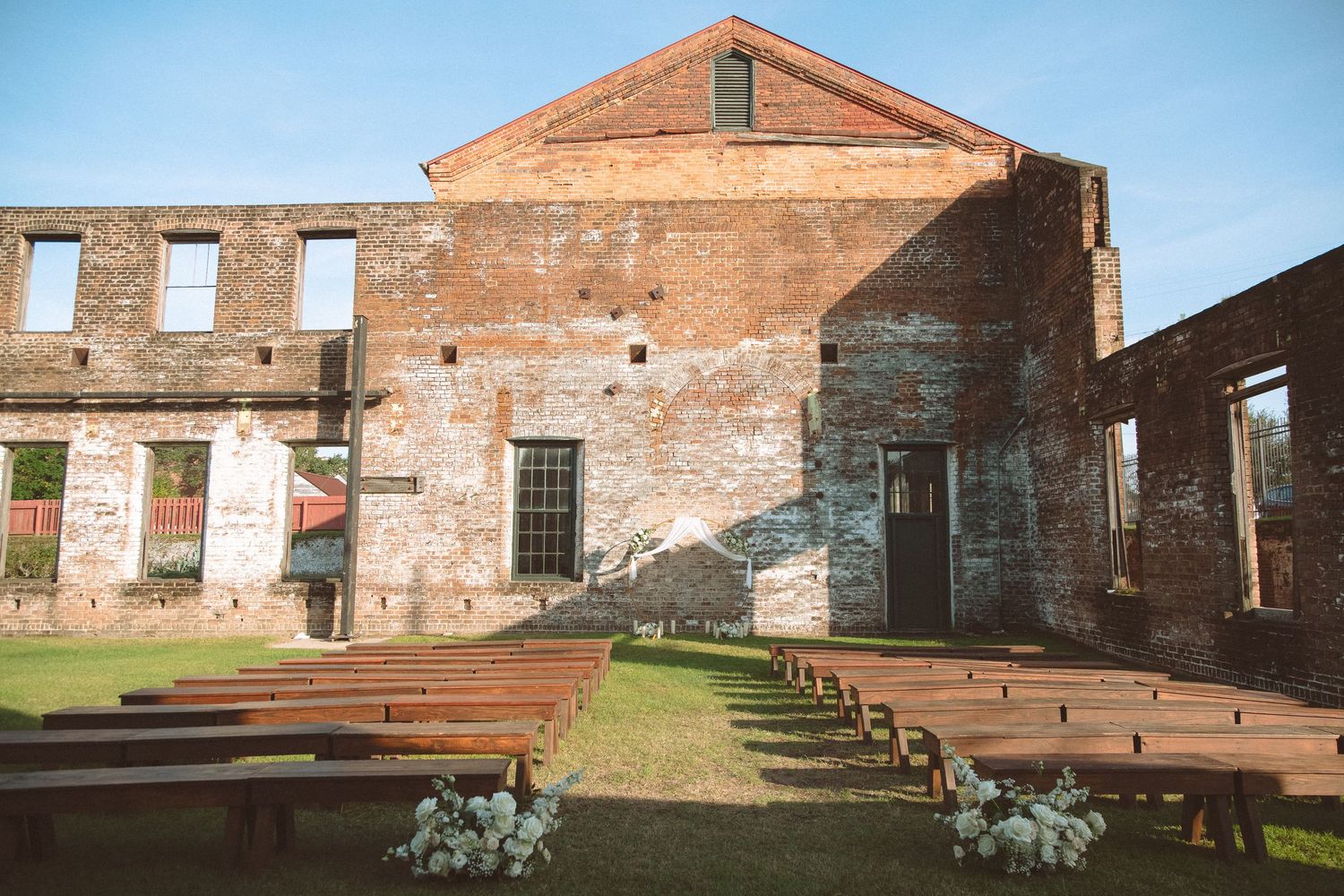 Outdoor wedding ceremony setup with wooden pews inside historic brick ruins on a sunny day.