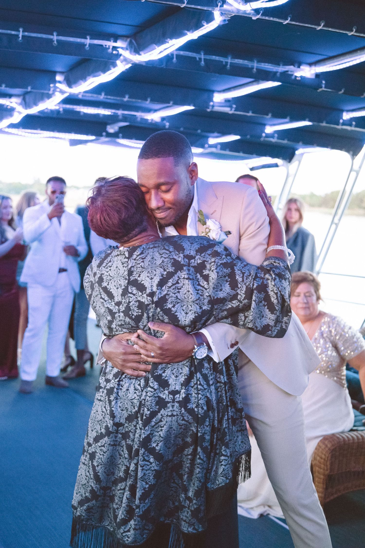 Groom in tan suit shares emotional dance with mother in silver dress at wedding.