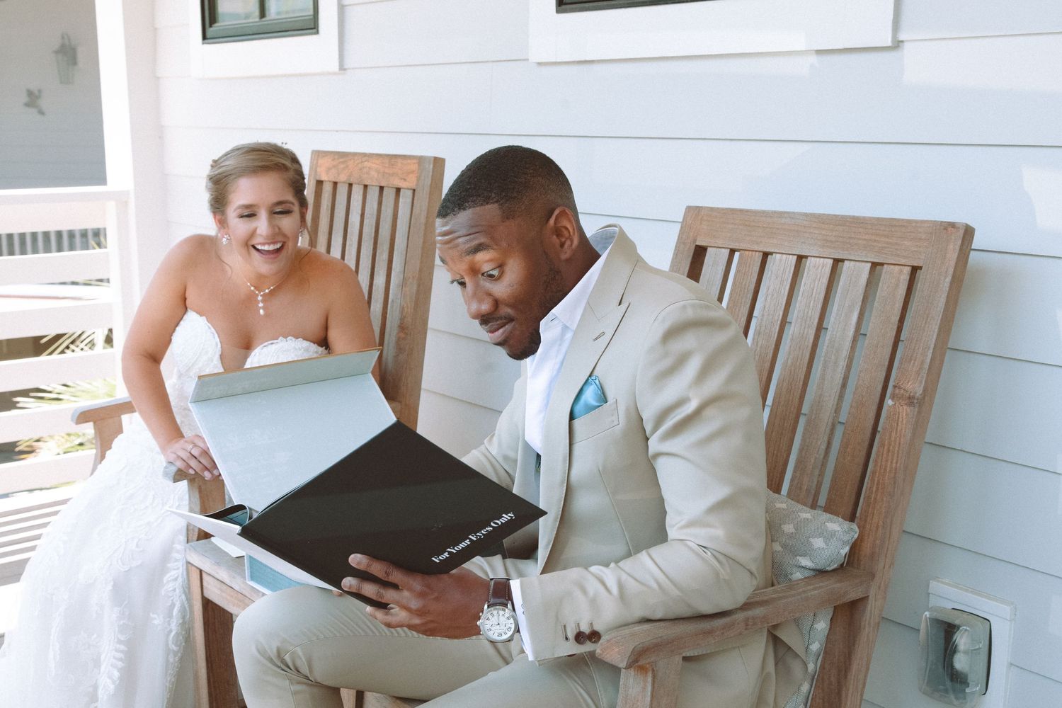 Bride smiling as groom in beige suit views their wedding photo album on a sunny porch.