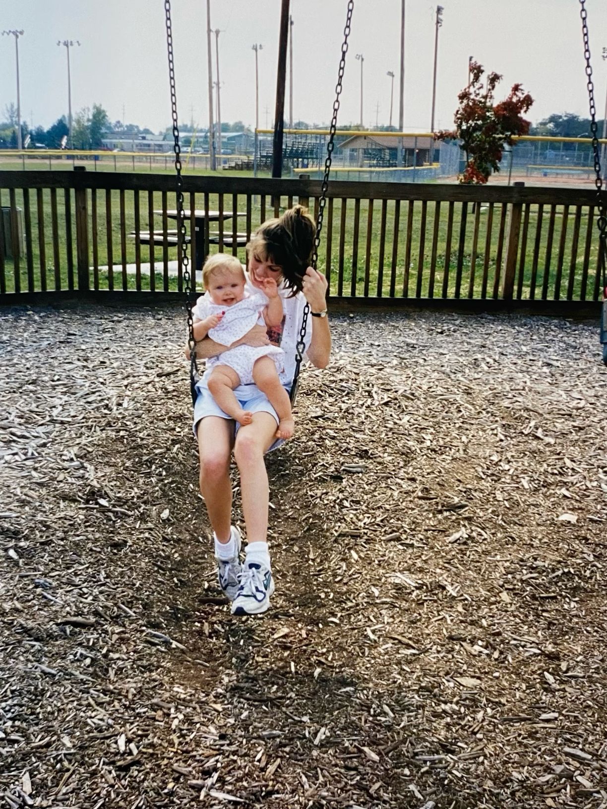 Mother and baby sitting together on a playground swing surrounded by wood chip ground cover.