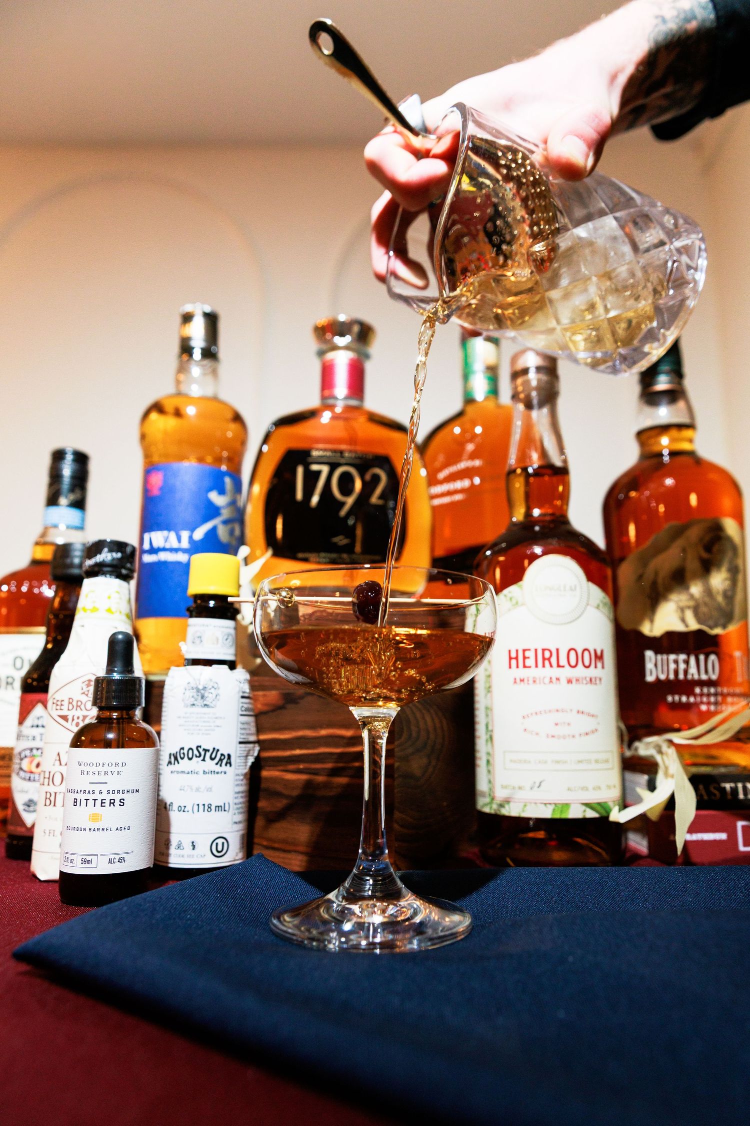 Bartender pouring whiskey cocktail into coupe glass with 1792 and Buffalo Trace bottles in background.
