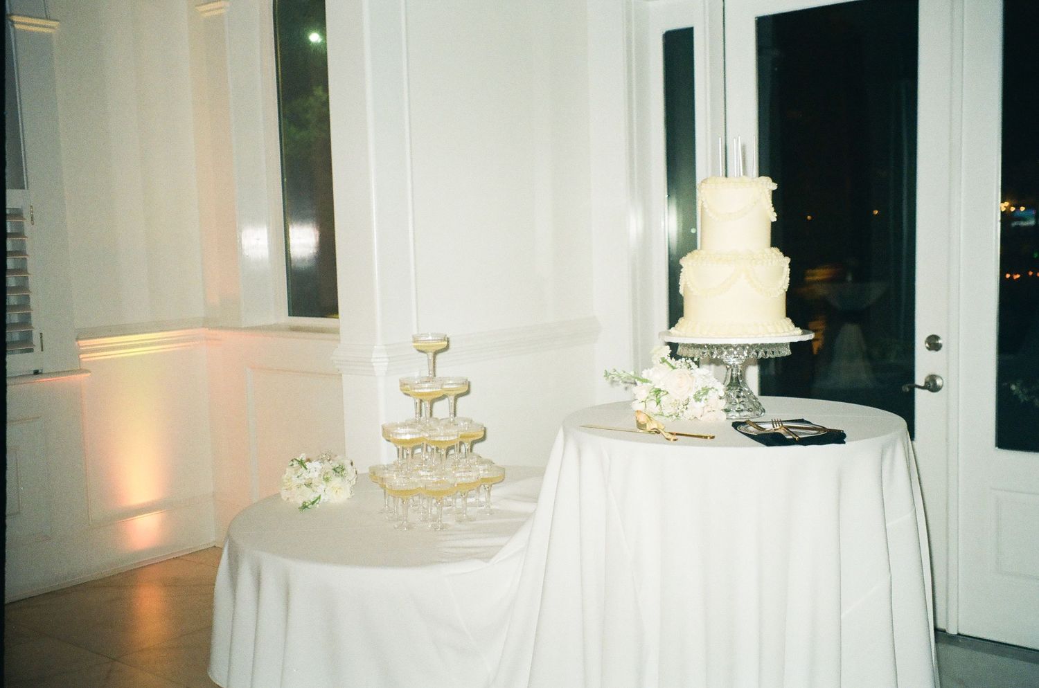 Elegant wedding reception display with a white tiered cake and champagne fountain tower on draped tables in a softly lit venue.