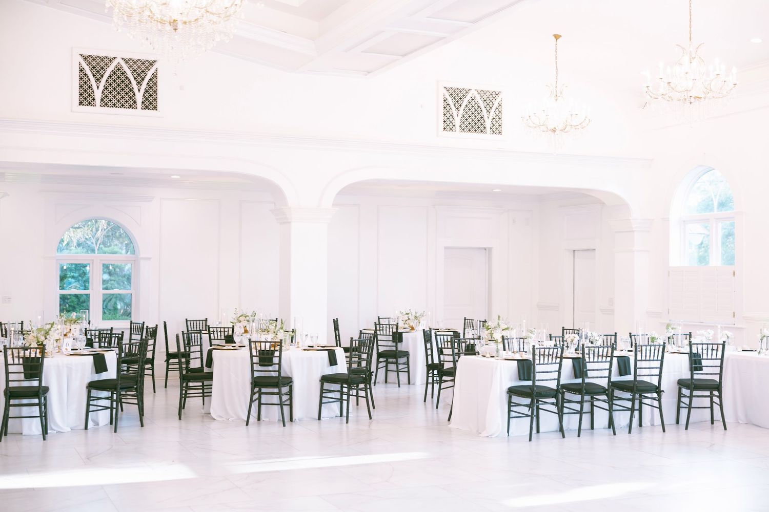 Elegant white ballroom with round tables, dark chiavari chairs, and chandelier lighting for a wedding reception.