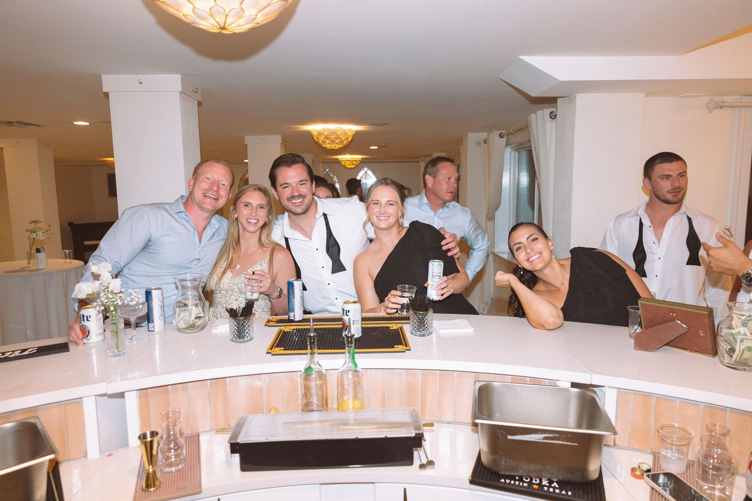 Group of smiling guests posing together at a bar during a wedding reception celebration.