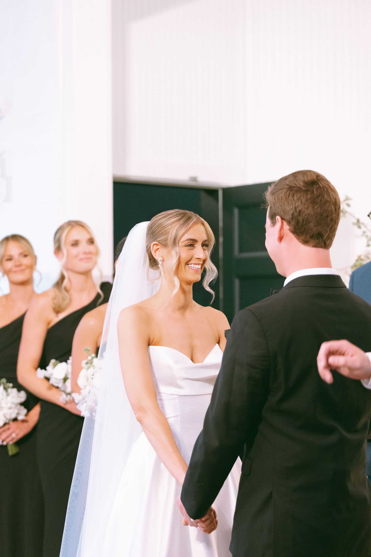 Smiling bride in white gown holds grooms hands at altar with bridesmaids in black dresses during wedding ceremony.