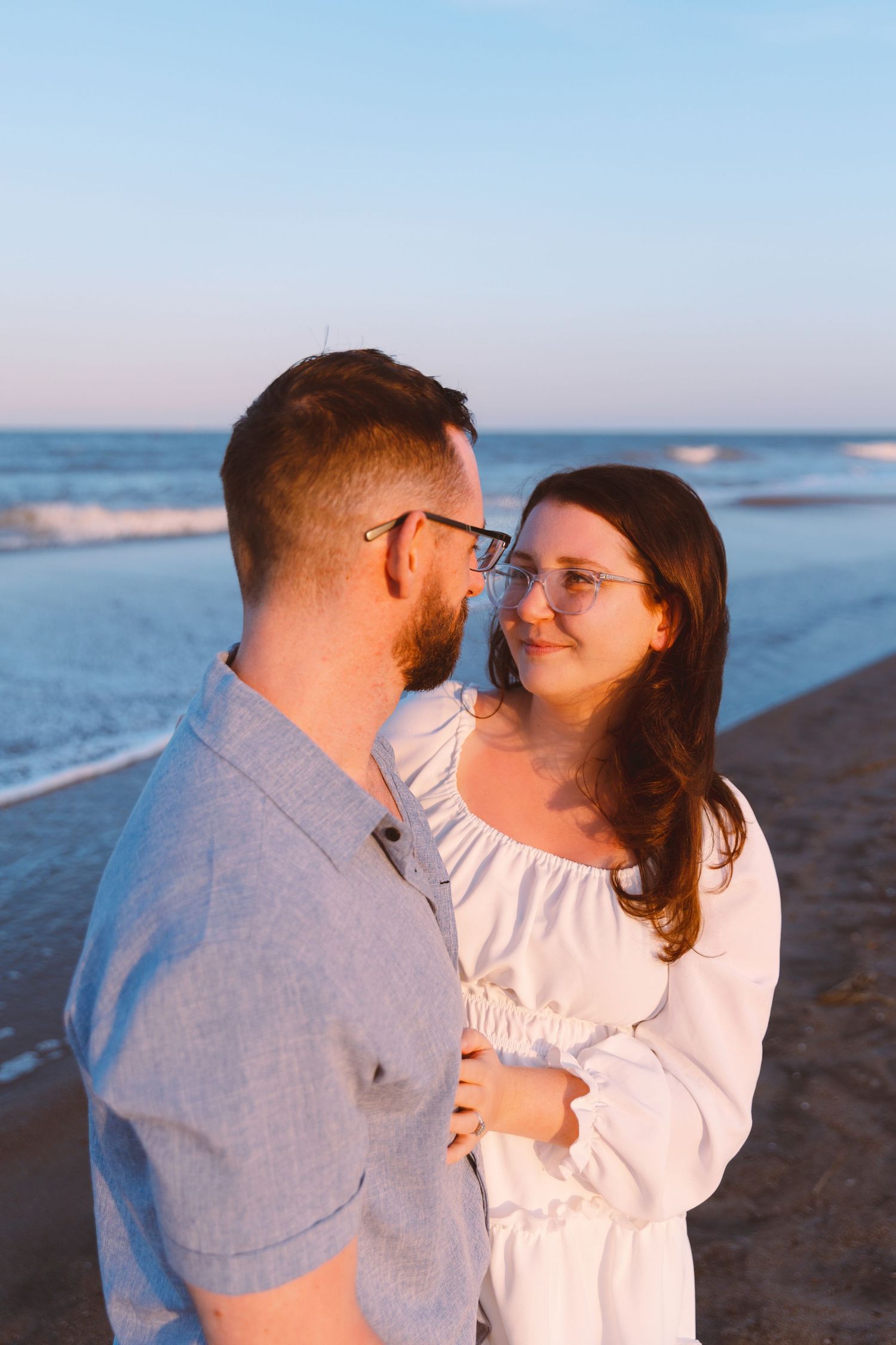 Couple embracing on a beach at sunset, woman in white dress and glasses smiling at man in gray shirt.