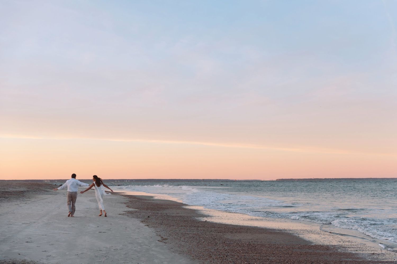 Couple holding hands walking along a sandy beach at sunset with soft pink and blue sky.