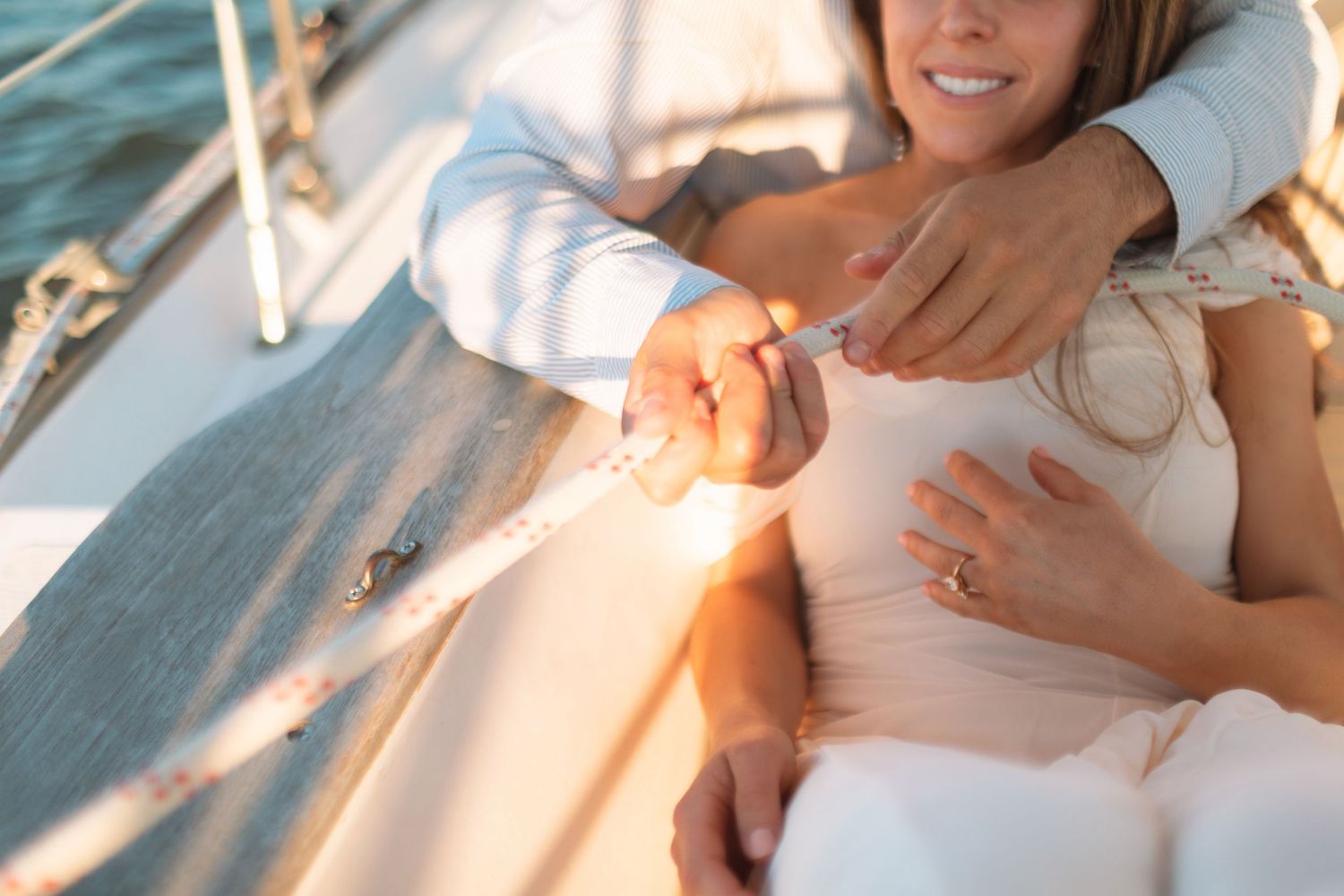 Couple relaxing on a sailboat deck, woman wearing engagement ring while partner embraces her in golden sunlight.