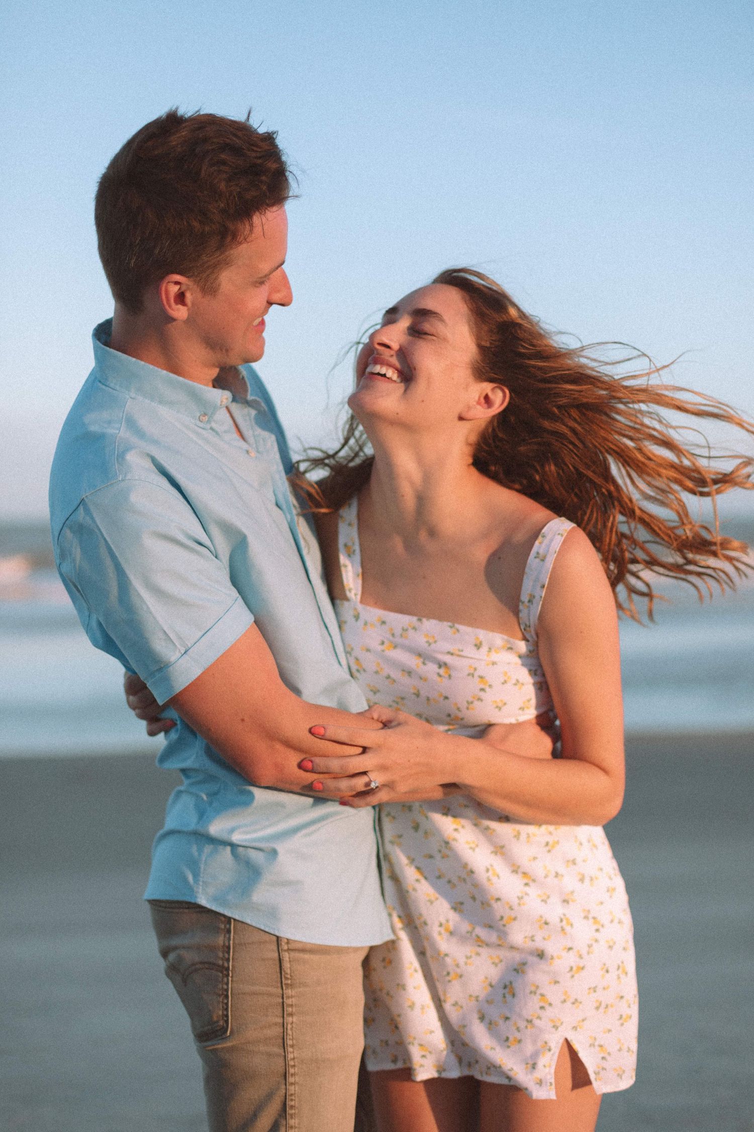 Happy couple embracing and laughing on a sunny beach, womans hair blowing in the wind.