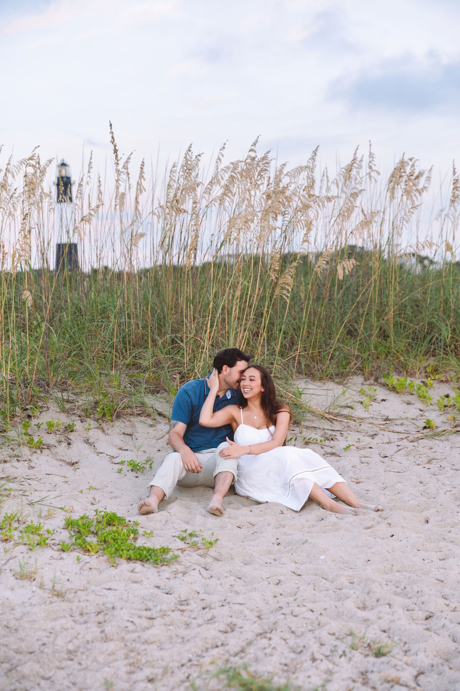 Couple sitting on sandy beach dunes with sea oats and lighthouse, man kissing woman in white dress.