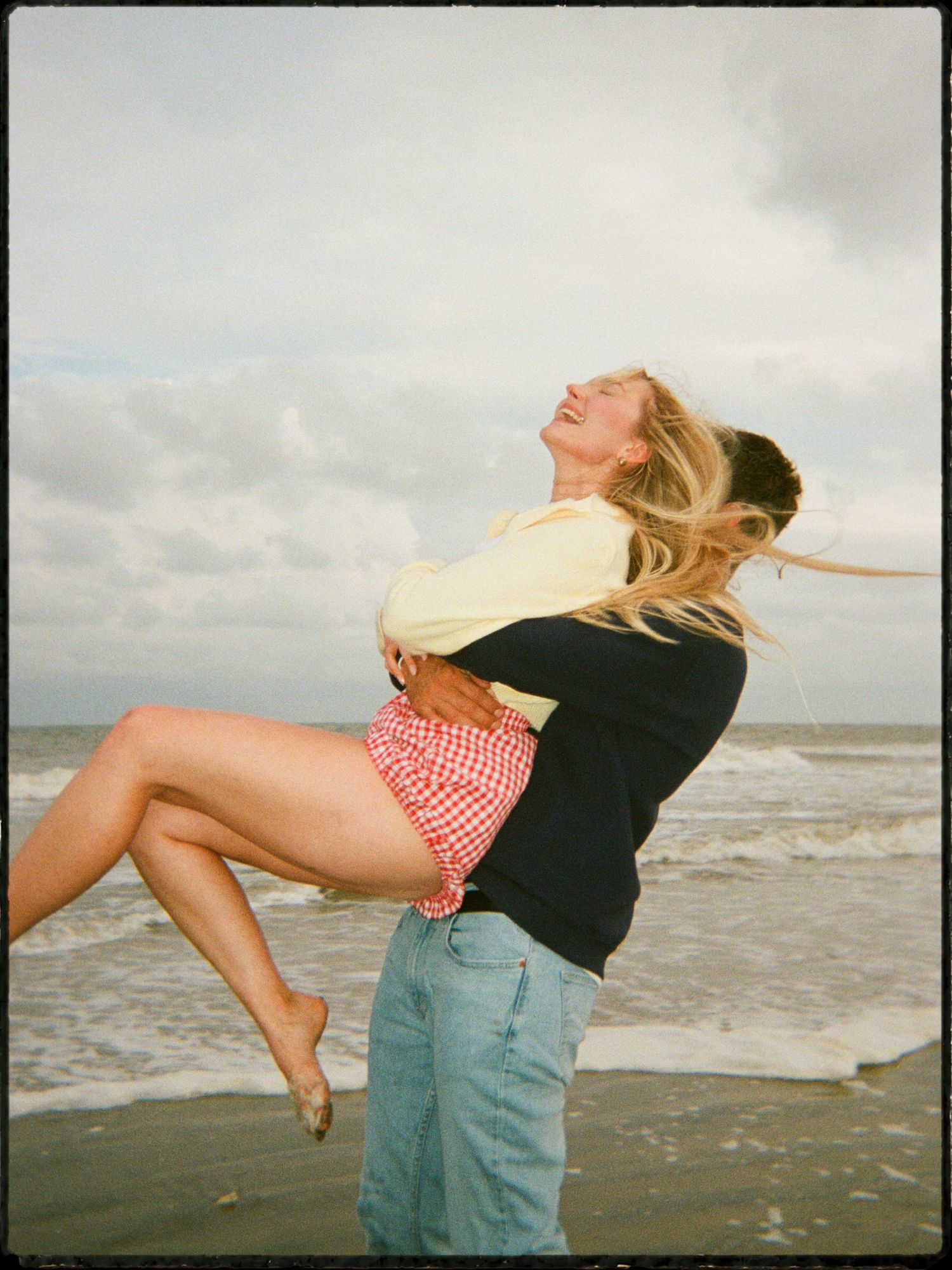 Man lifting and spinning a laughing woman on a windy beach, with ocean waves and cloudy skies in the background.