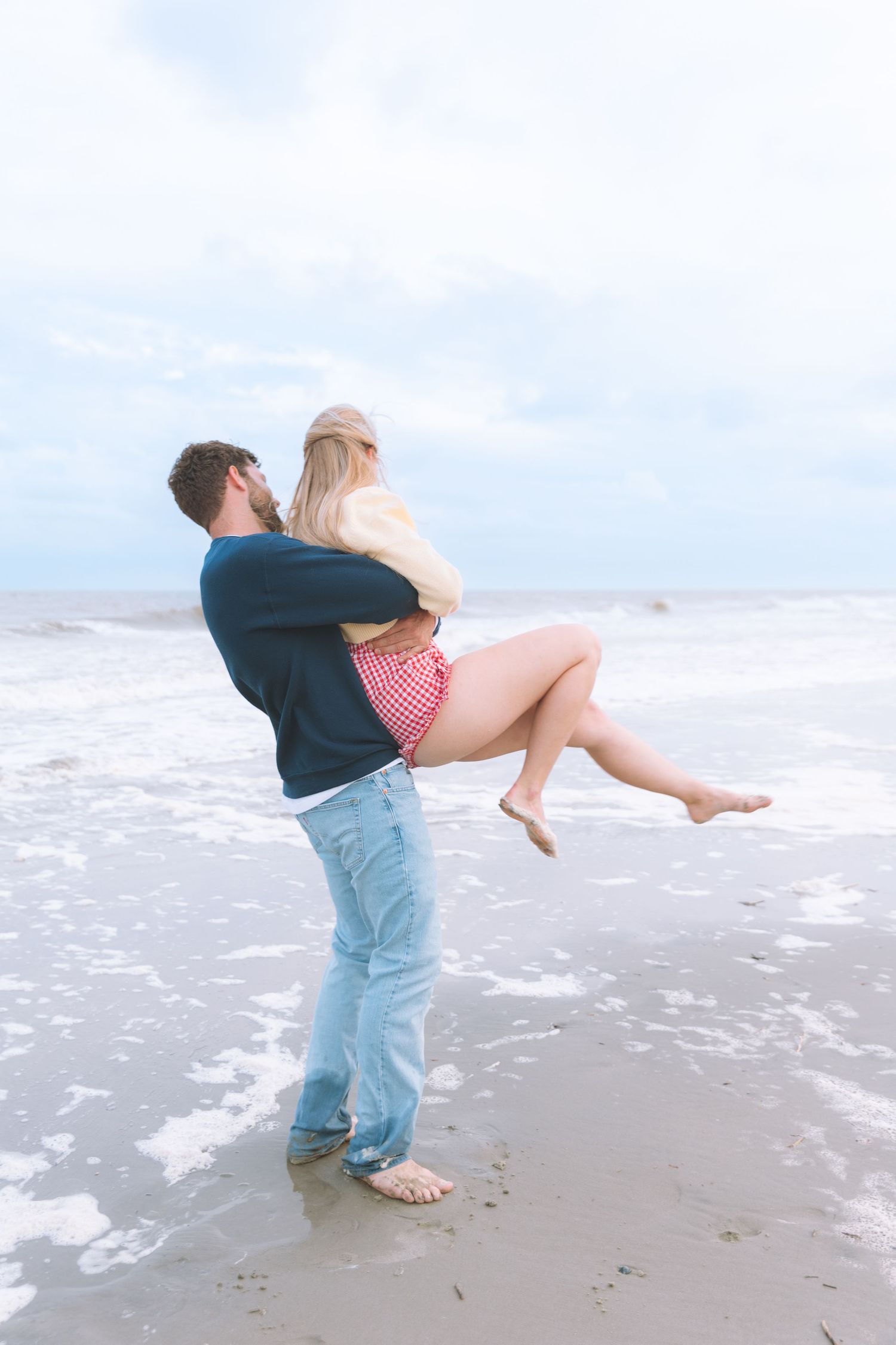 Man lifting woman in his arms while standing in ocean waves on a beach.