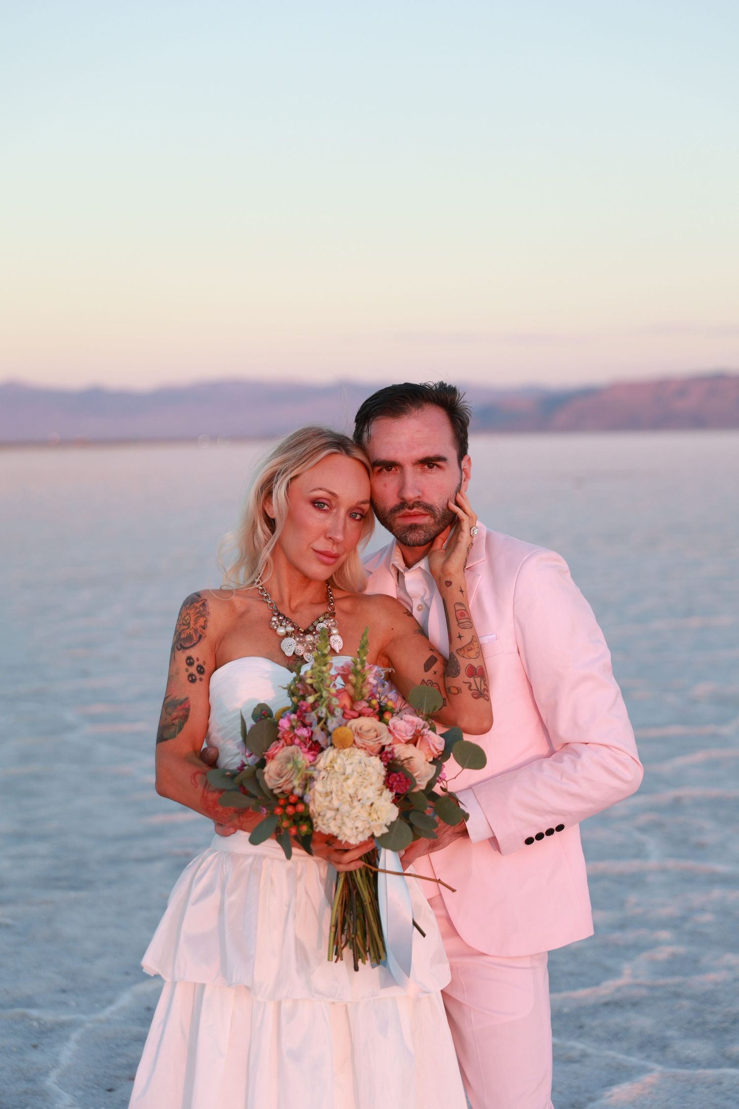 Tattooed bride and groom in pink suit pose with floral bouquet at sunset on salt flats.