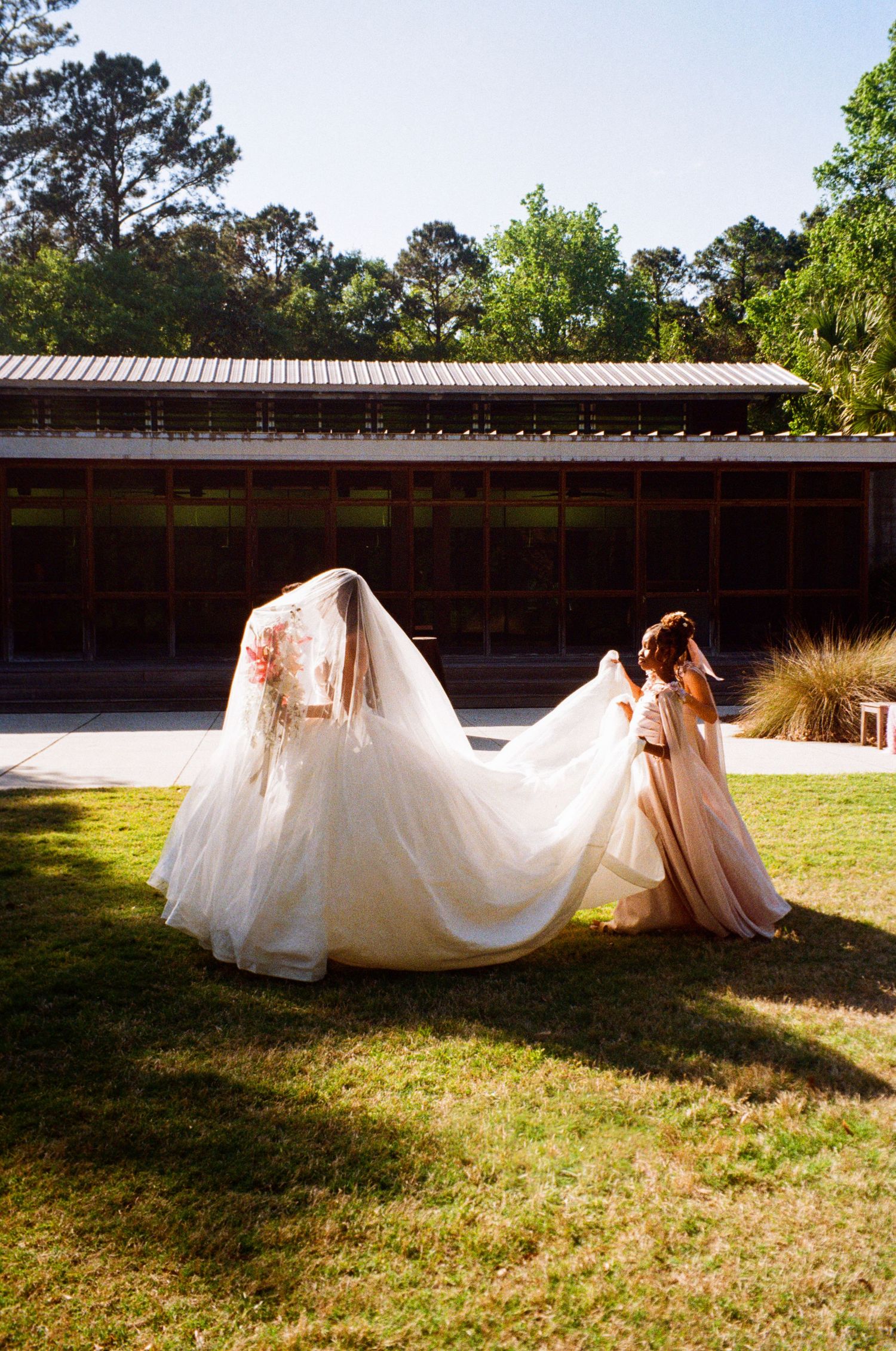 Bride and bridesmaid in blush dress walking outdoors with flowing white wedding gown and veil in sunlight.