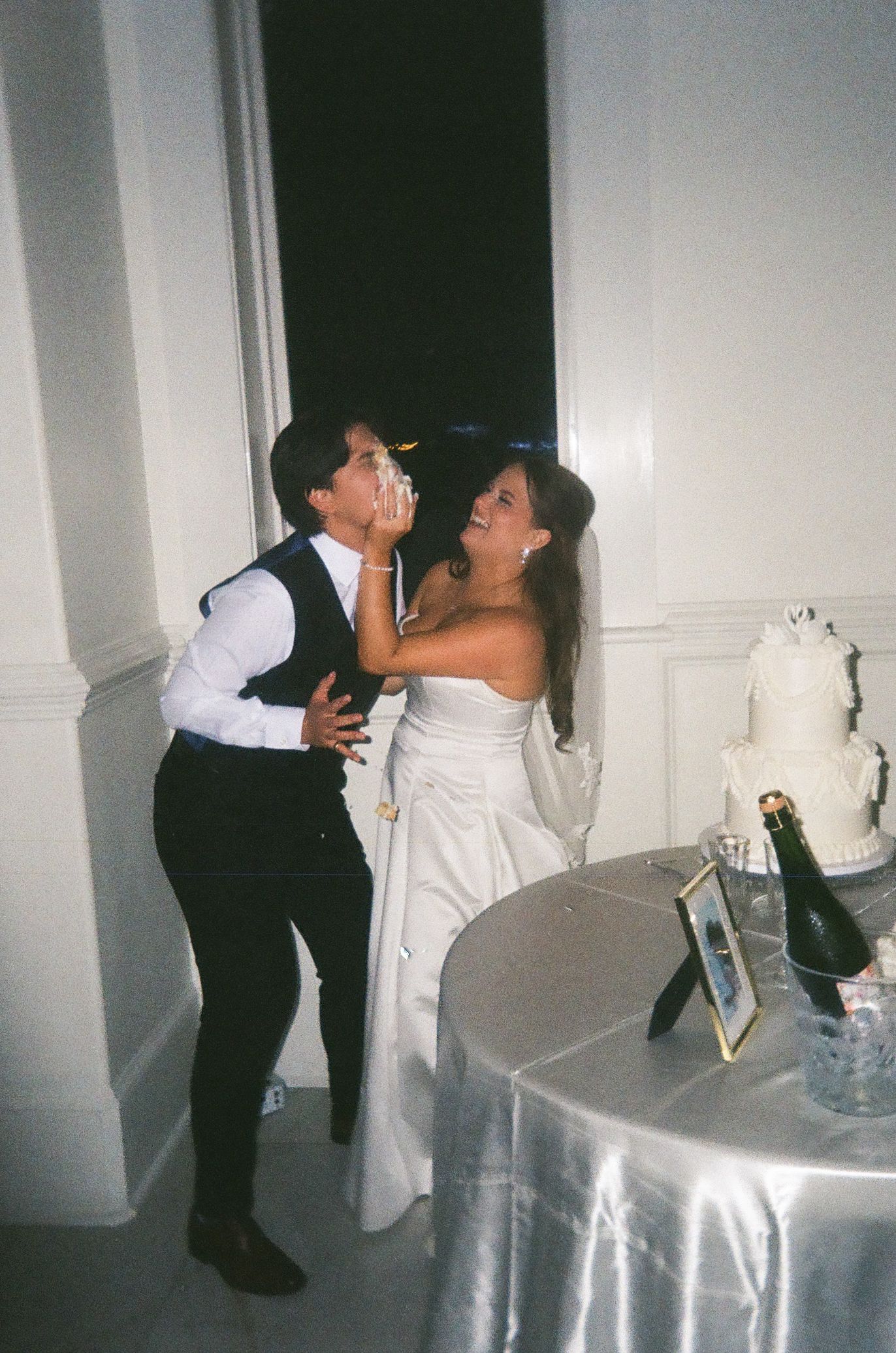 Bride in white gown playfully feeds cake to groom in black vest at elegant wedding reception.